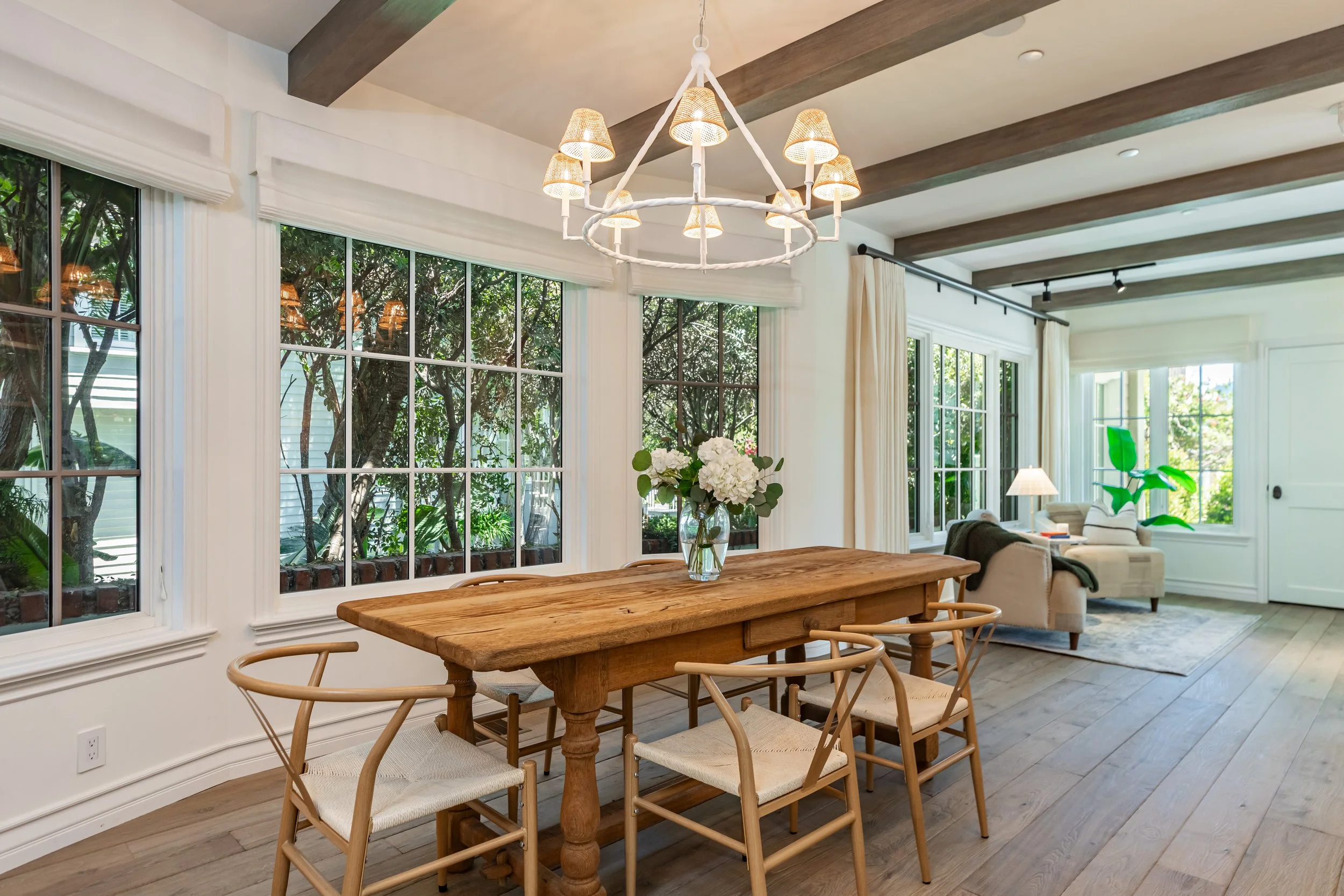 A dining room with a wooden table and beige chairs, large windows with white blinds, a modern chandelier, and a view into a living room with white walls, white curtains, and a green plant.