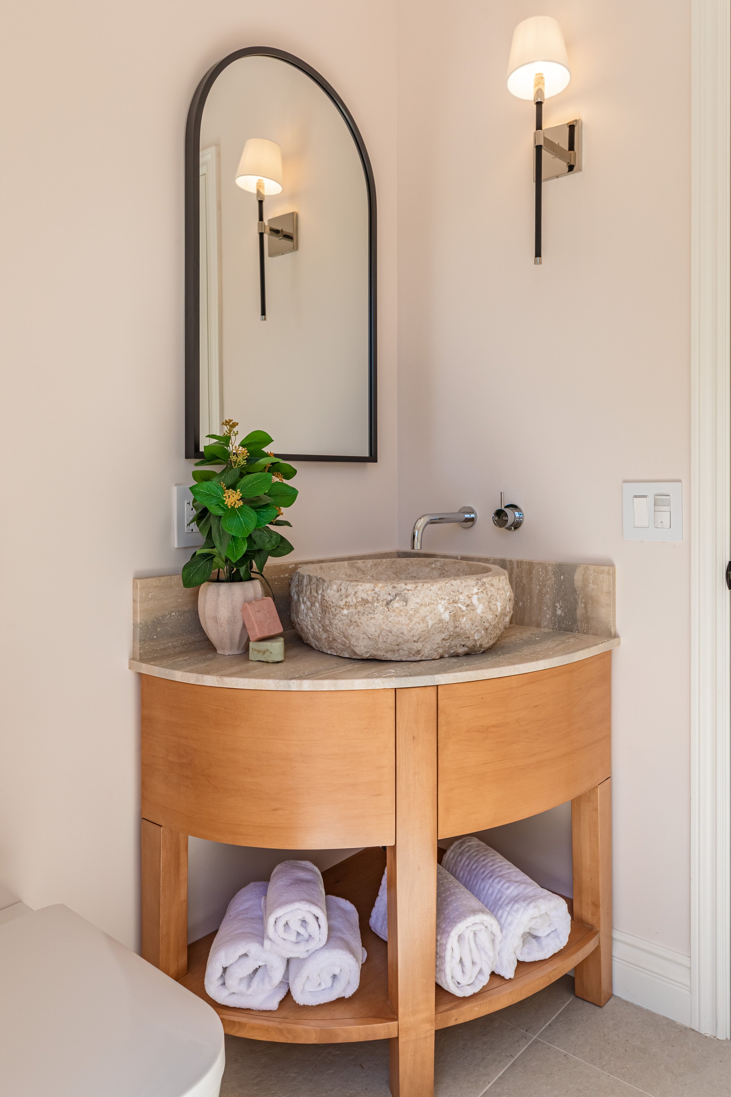 A bathroom vanity with a curved wooden cabinet, a stone vessel sink, and a round mirror. Blue and white rolled towels are stored underneath. A potted green plant with yellow flowers is on the counter, with a pink soap bar and a small stone beside it.