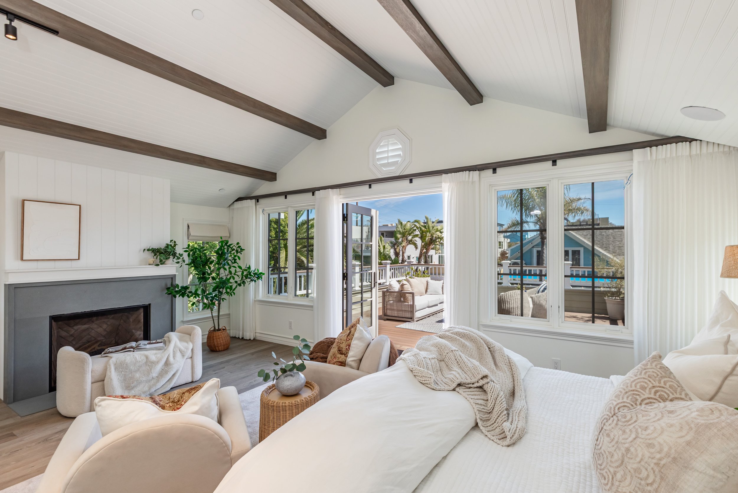 Living room with white walls, a fireplace, and large windows opening to a balcony with outdoor seating and palm trees.
