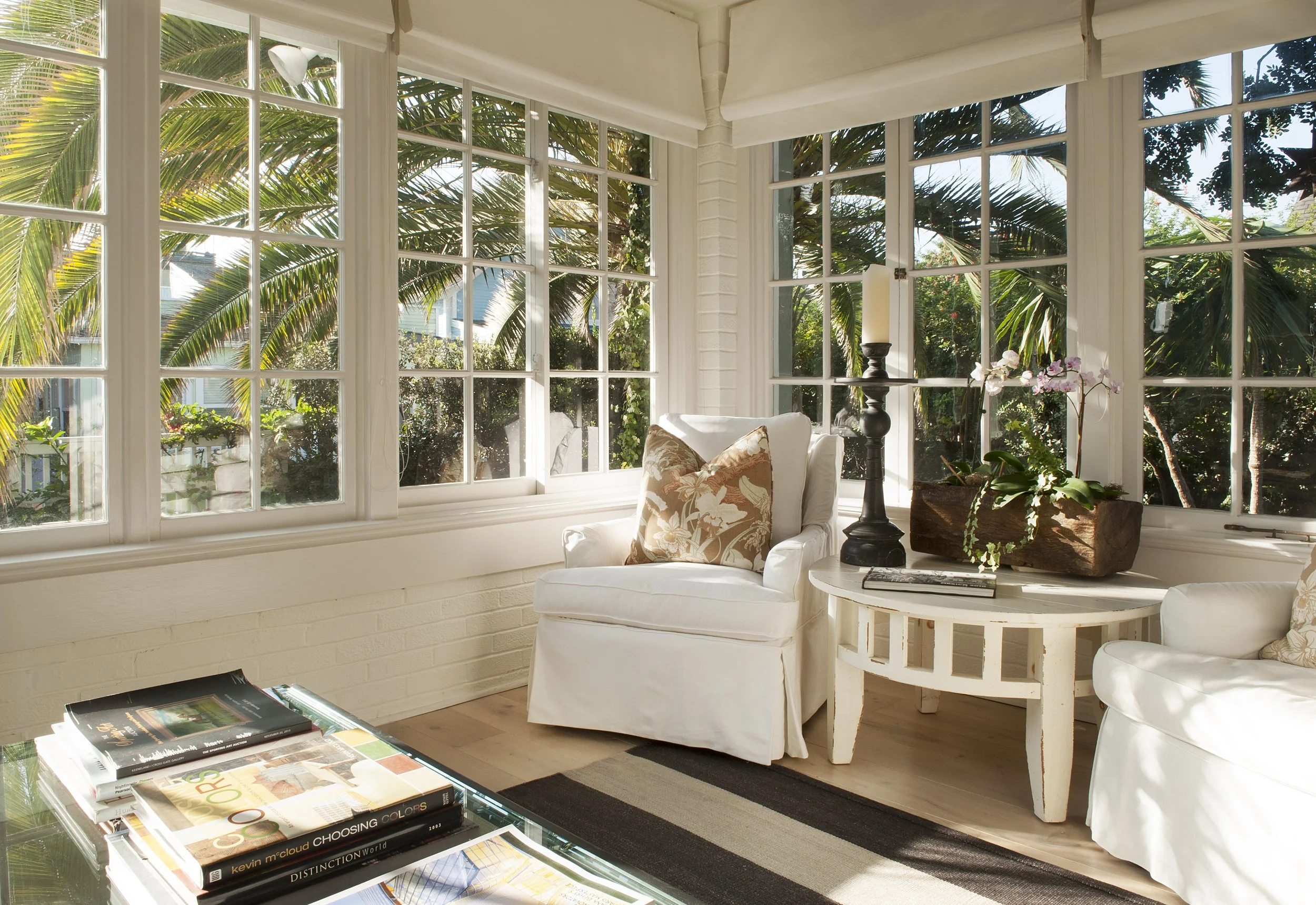 A sunlit room with large windows showing lush green palm trees outside. Inside, there is a white armchair with floral cushions, a small side table with a potted orchid and a candle holder, and a glass coffee table stacked with books.