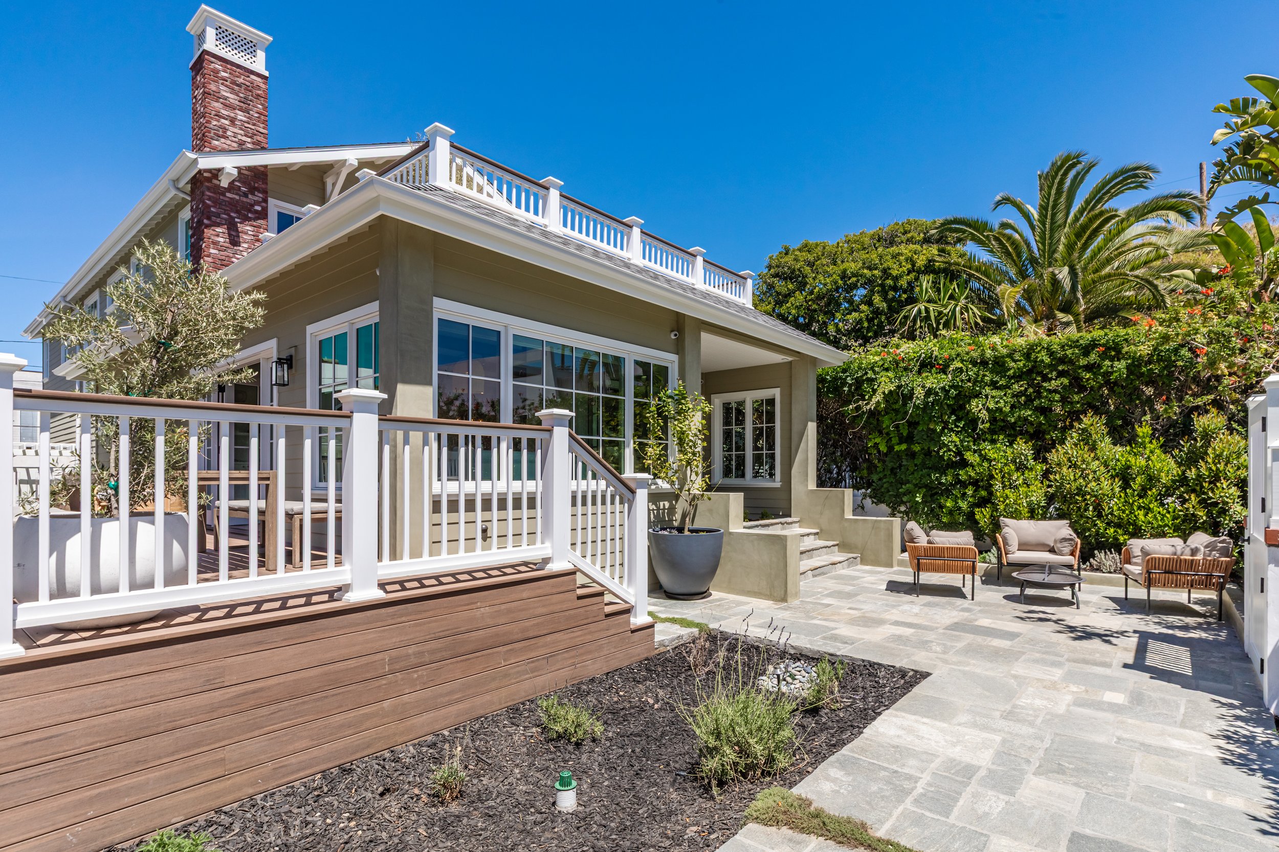 A modern house with a large outdoor patio area featuring seating, a wooden deck, potted plants, and a garden with palm trees under a clear blue sky.