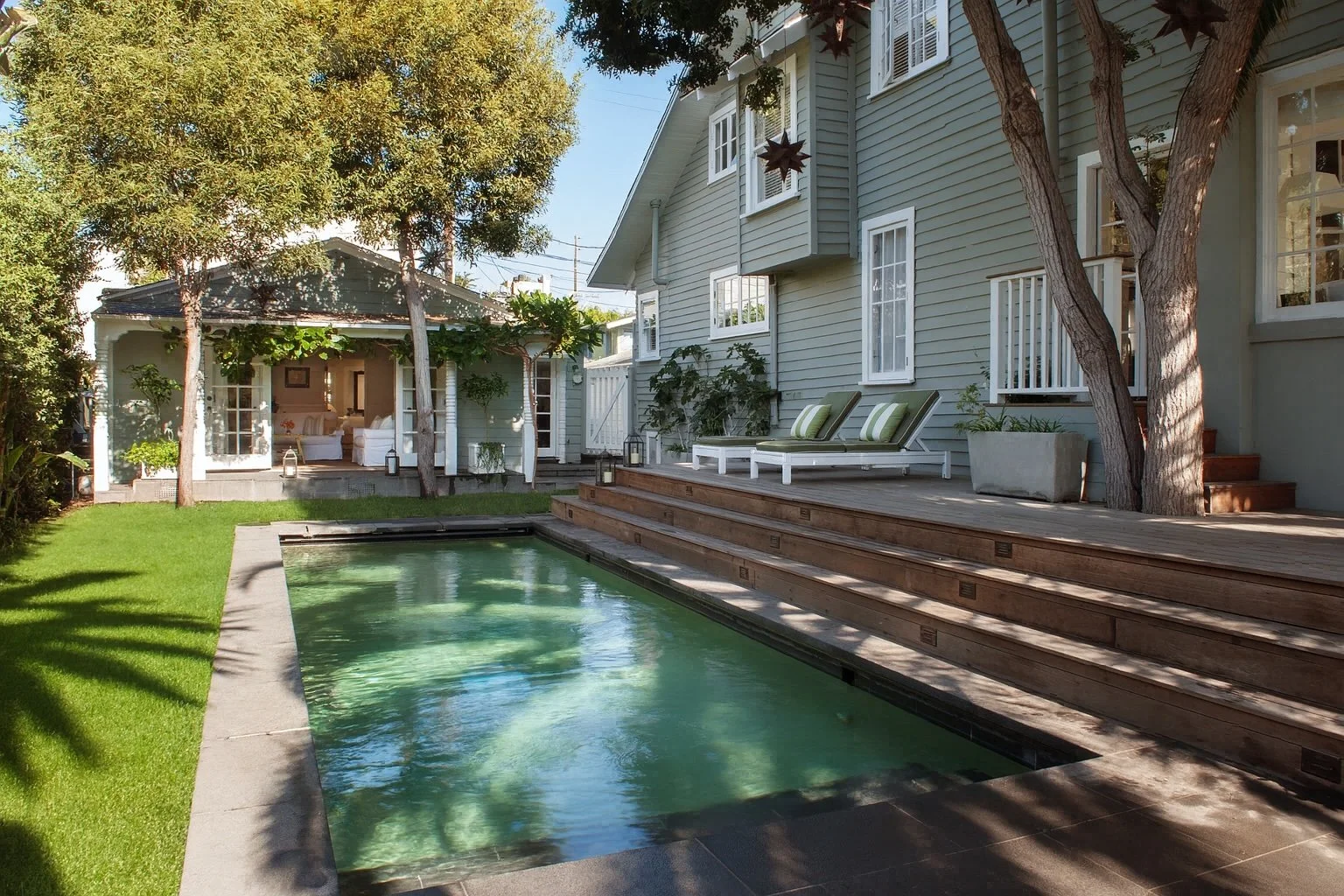 Backyard with a swimming pool, wooden deck with lounge chairs, outside house with large windows, patio furniture in a screened porch, lush green lawn, trees, and potted plants.