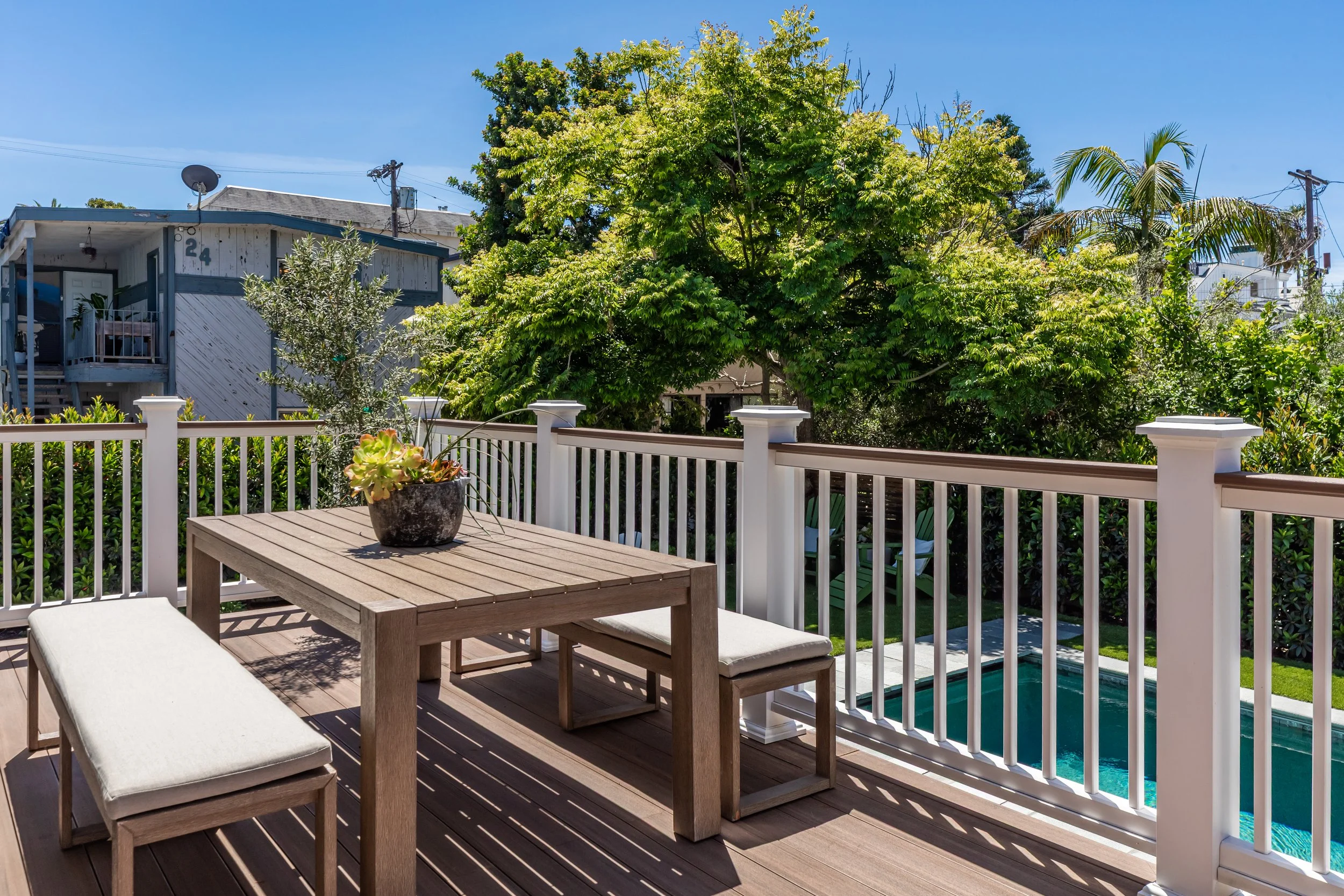 A backyard patio with a wooden dining table and cushioned benches, surrounded by a white fence, overlooking a green garden with large trees and a swimming pool.