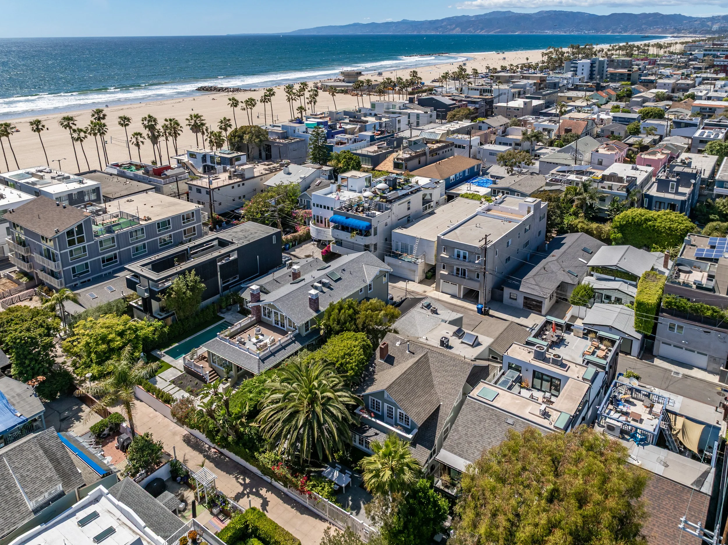 Aerial view of a coastal neighborhood with houses and palm trees near the beach and ocean, with mountains in the background.