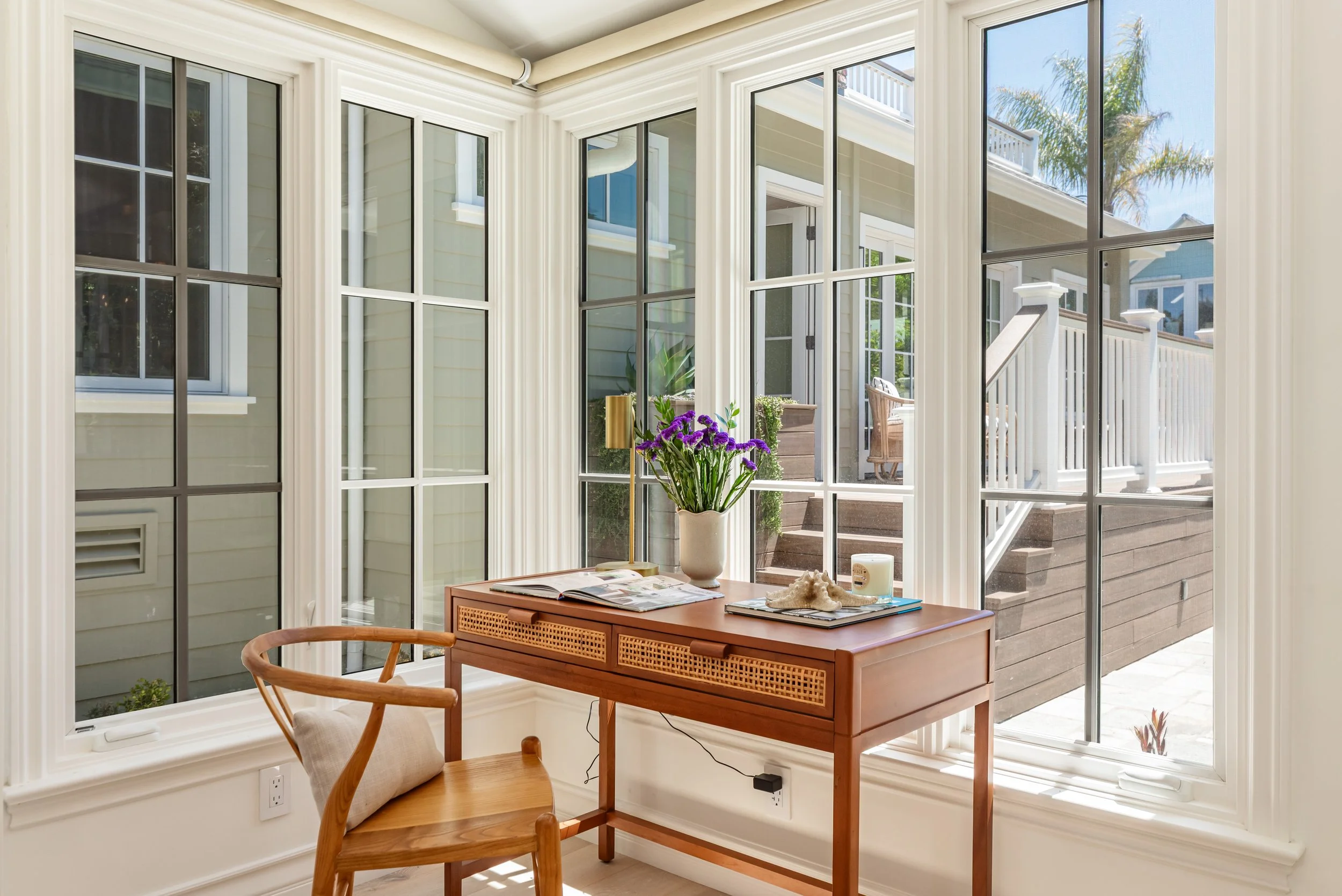 Sunlit interior room with large window and sliding glass door looking out onto a deck and palm trees. There is a wooden table with books, a shell, and a potted purple flower, along with a wooden chair.