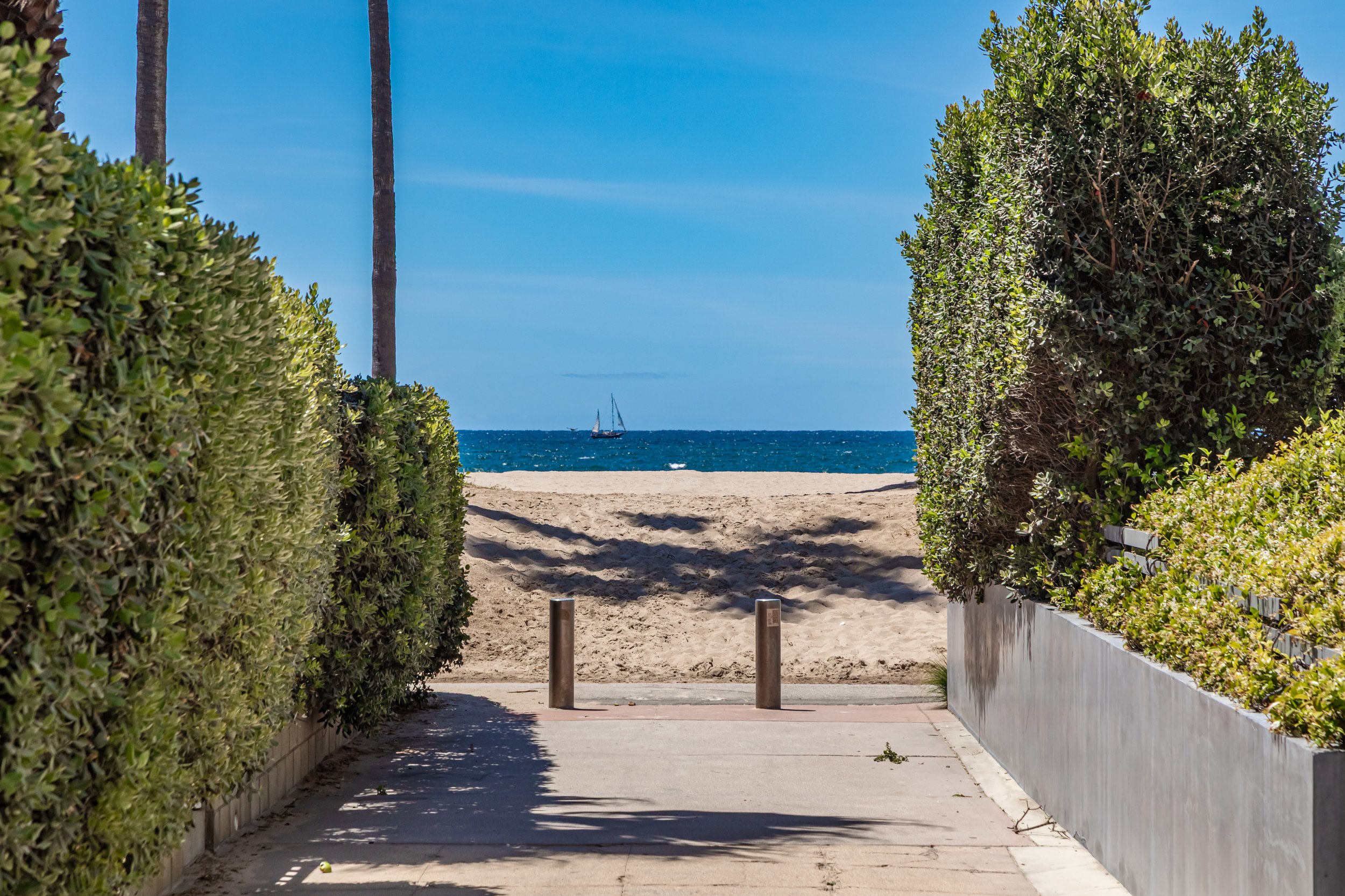 A pathway leading to a beach with the ocean and a sailboat visible in the distance. The path is bordered by green bushes and trees, with a clear blue sky above.