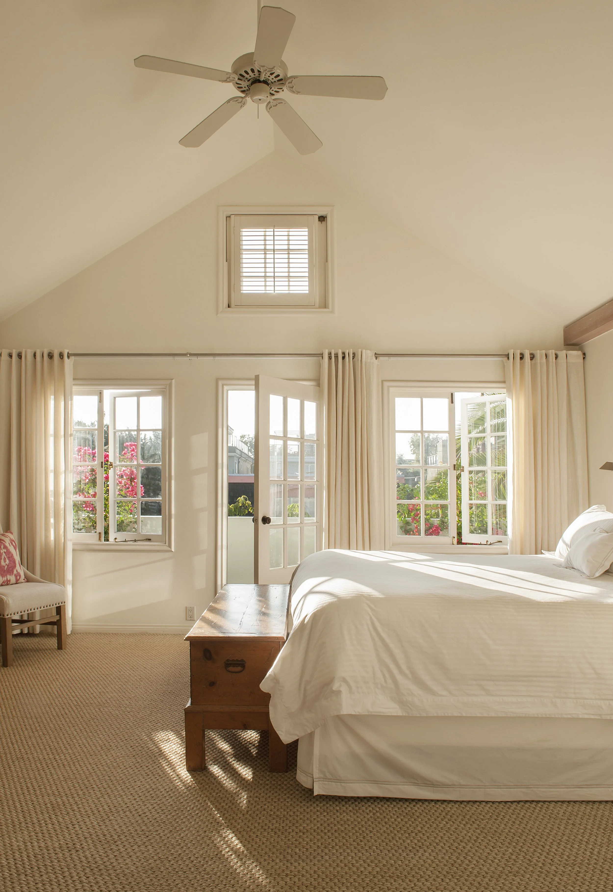 Sunlit bedroom with large windows, cream curtains, a white bed, a wooden chest at the foot of the bed, and a chair with a patterned cushion, ceiling fan, and garden view outside.