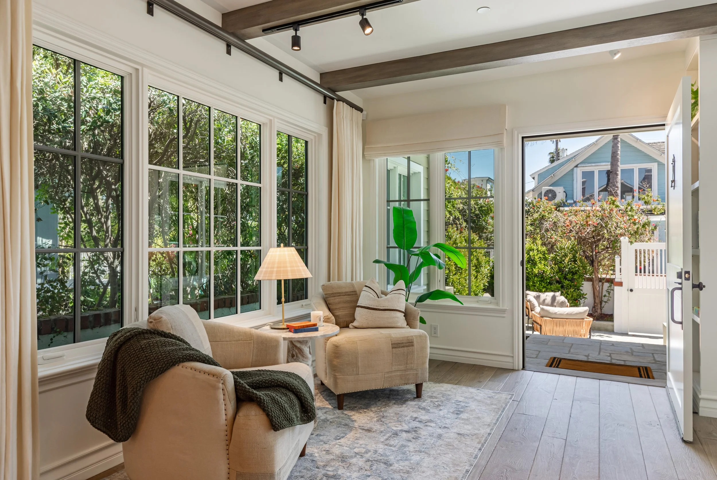 Bright living room with large windows showing lush greenery outside, beige armchairs with striped pillows, a small round table with books and a lamp, and a view of a backyard with a house, trees, and flowers.