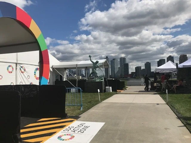 Outdoor event space with tents, a sculpture of a person with arms raised, and a city skyline in the background beneath a partly cloudy sky.