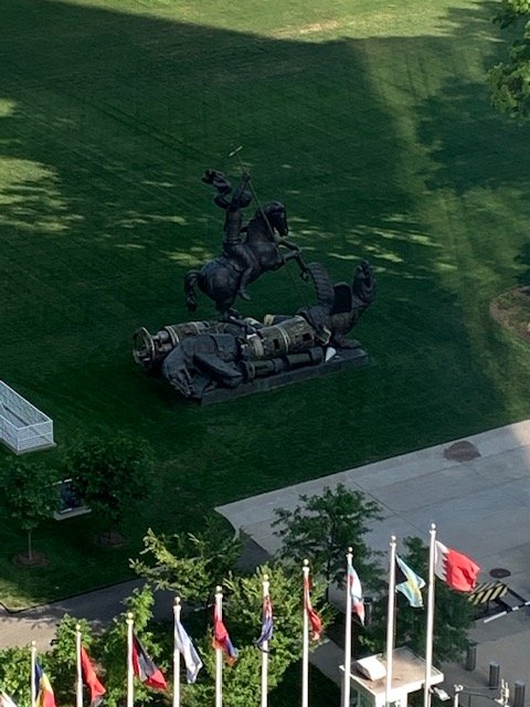 A large statue of a black horse rearing up, which appears to be a sculpture, is situated on a grassy area with trees, near a sidewalk with flags.