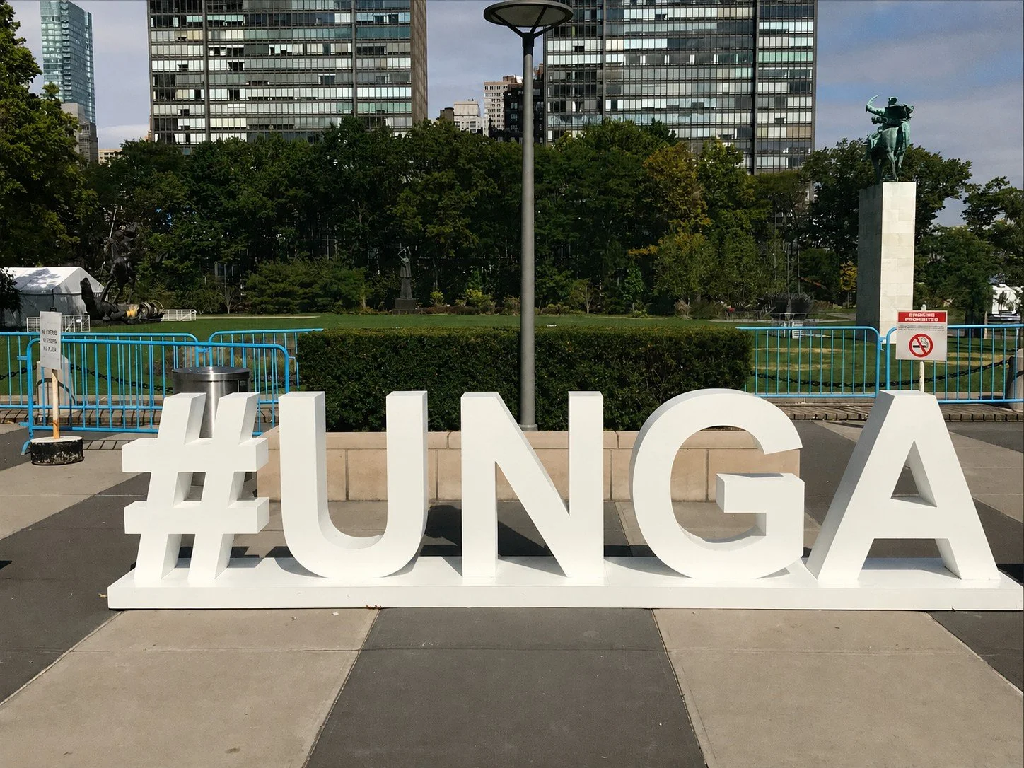 Large white hashtag sign with '#UNG' letters on a sidewalk in front of a park with green trees, sculptures, and modern glass buildings in the background.