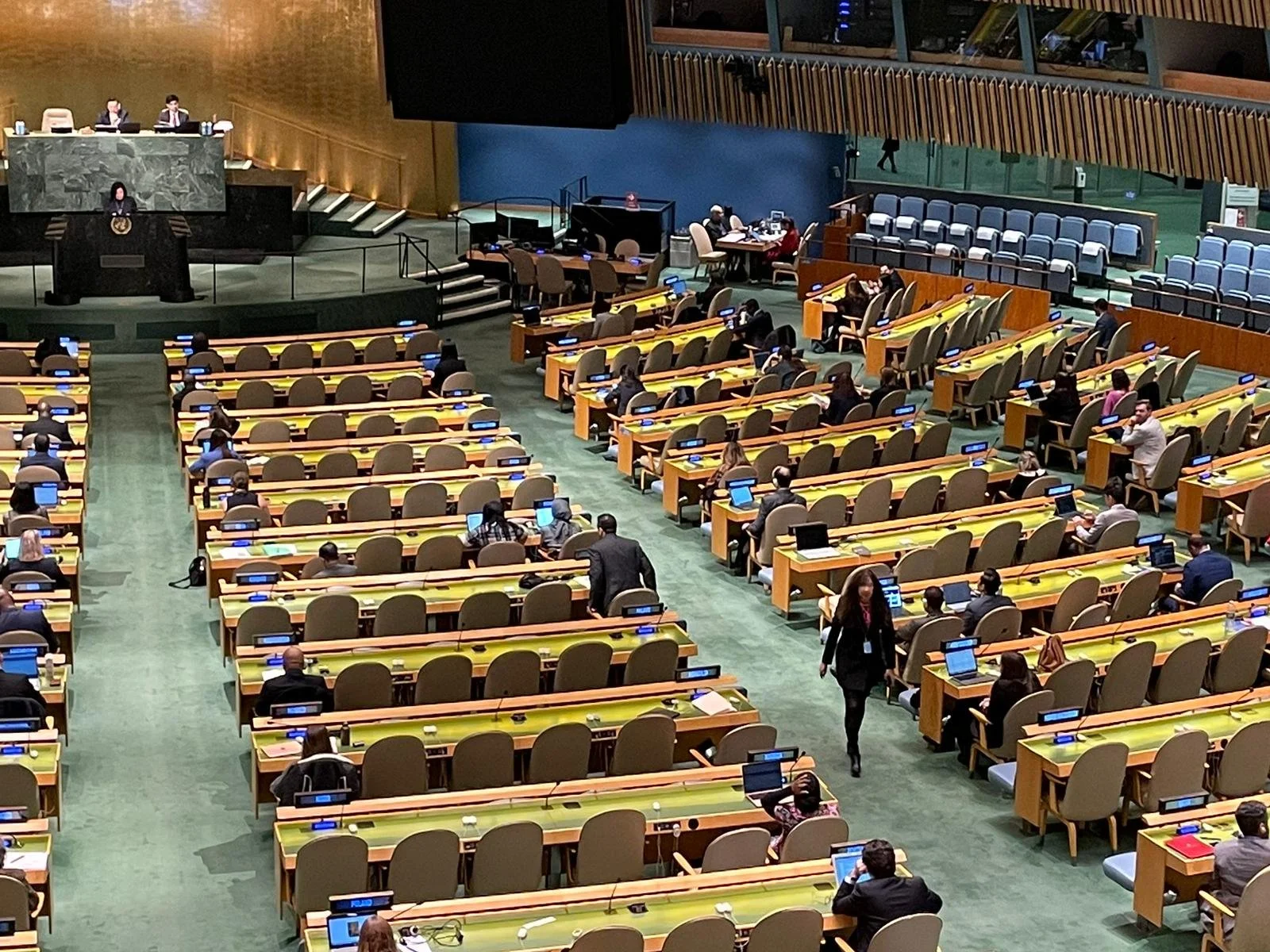 A large conference room or assembly hall with many attendees seated at desk stations with computers. The room is designed for a formal or international meeting, with a stage and podium at the front, and a person walking between the rows of desks.