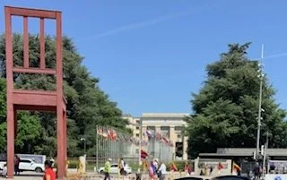 City street scene with trees, a building in the background, flags, parked cars, and people walking.