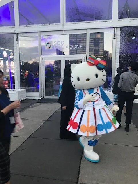 Hello Kitty mascot dressed in a colorful, rainbow-themed dress and bow, walking outside a glass building with people nearby.