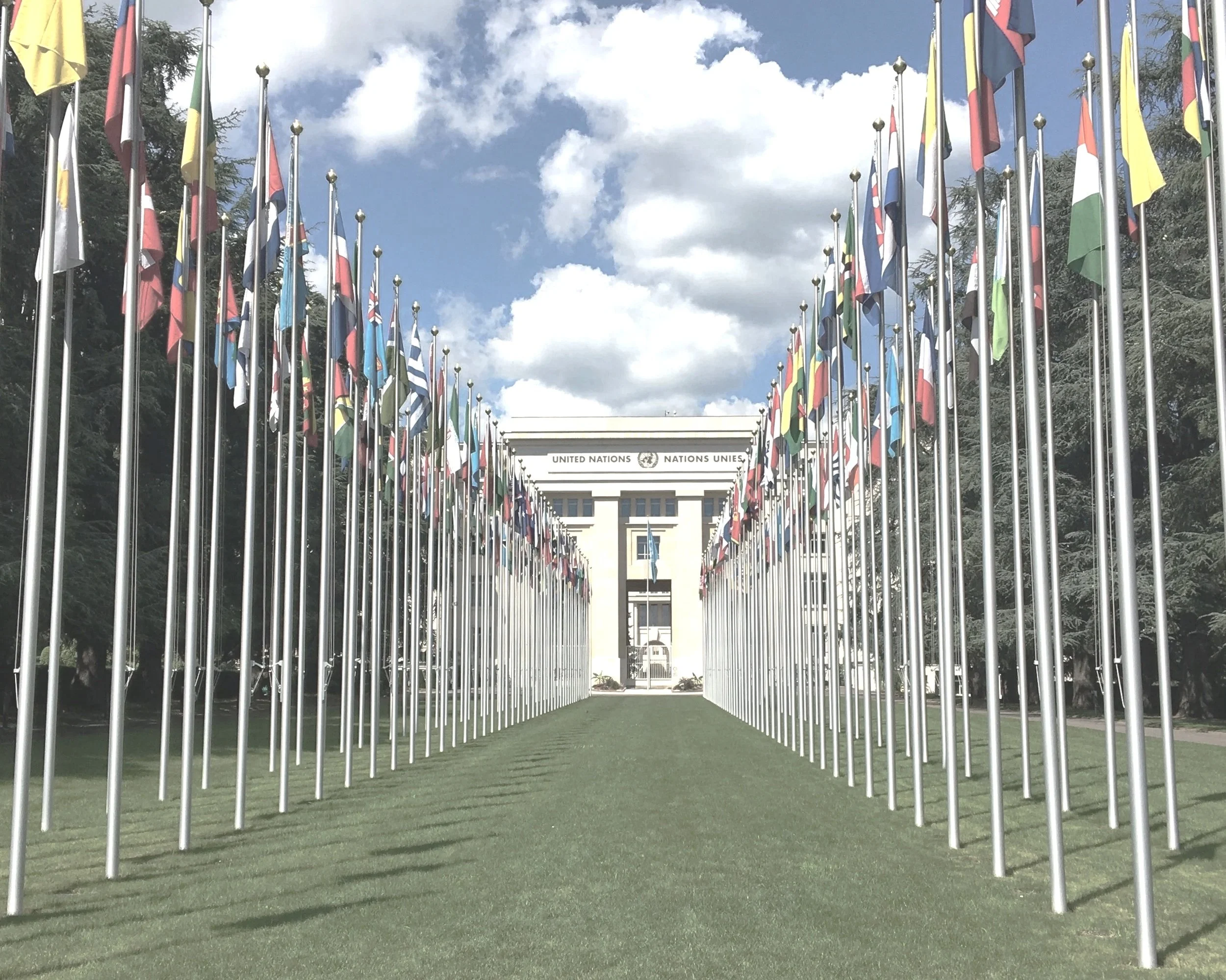Flagpoles with international flags surrounding the United Nations building under a partly cloudy sky.