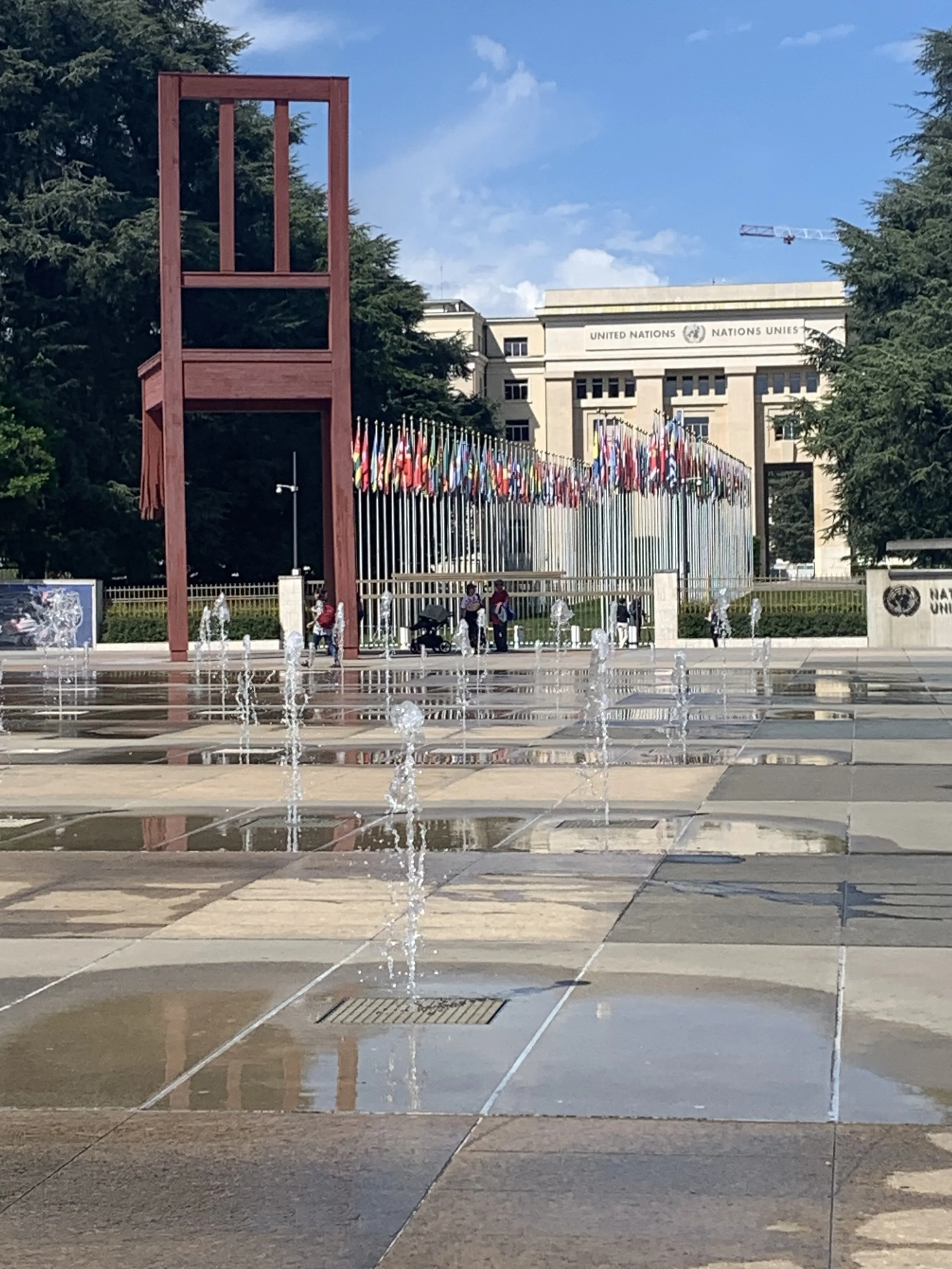 View of the United Nations headquarters with a fountain in the foreground, flags of various countries lined up along the fence, and a clear blue sky.