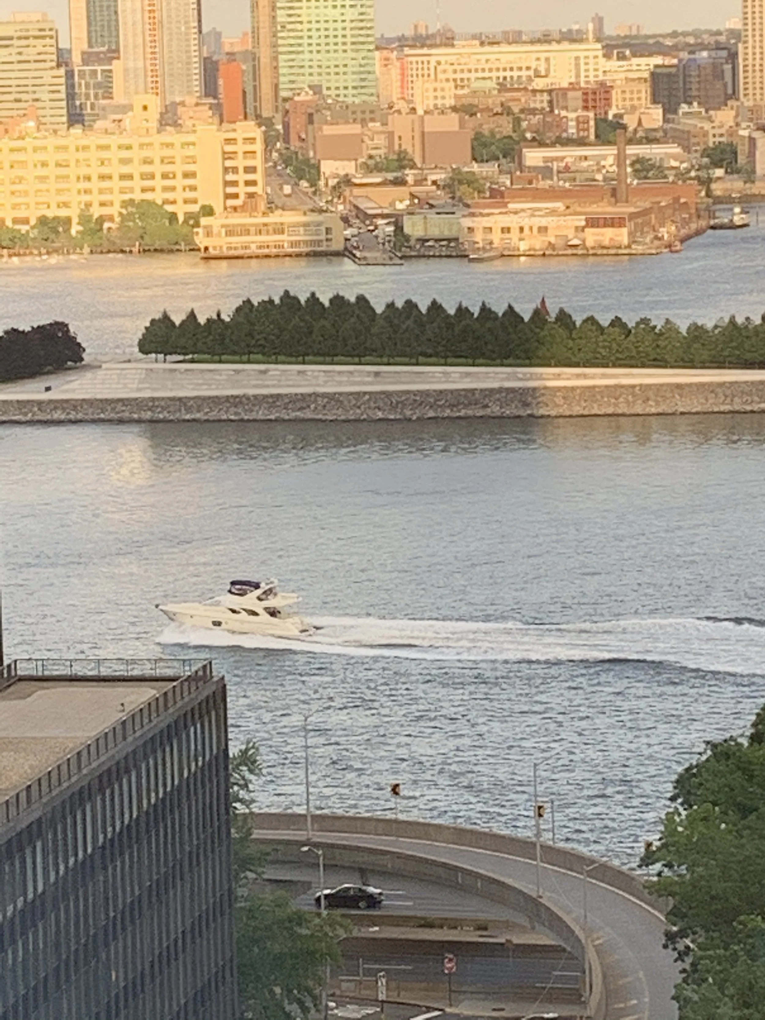 Cityscape with skyscrapers across a river, a yacht in the water, and a road with a car and a tunnel in the foreground.