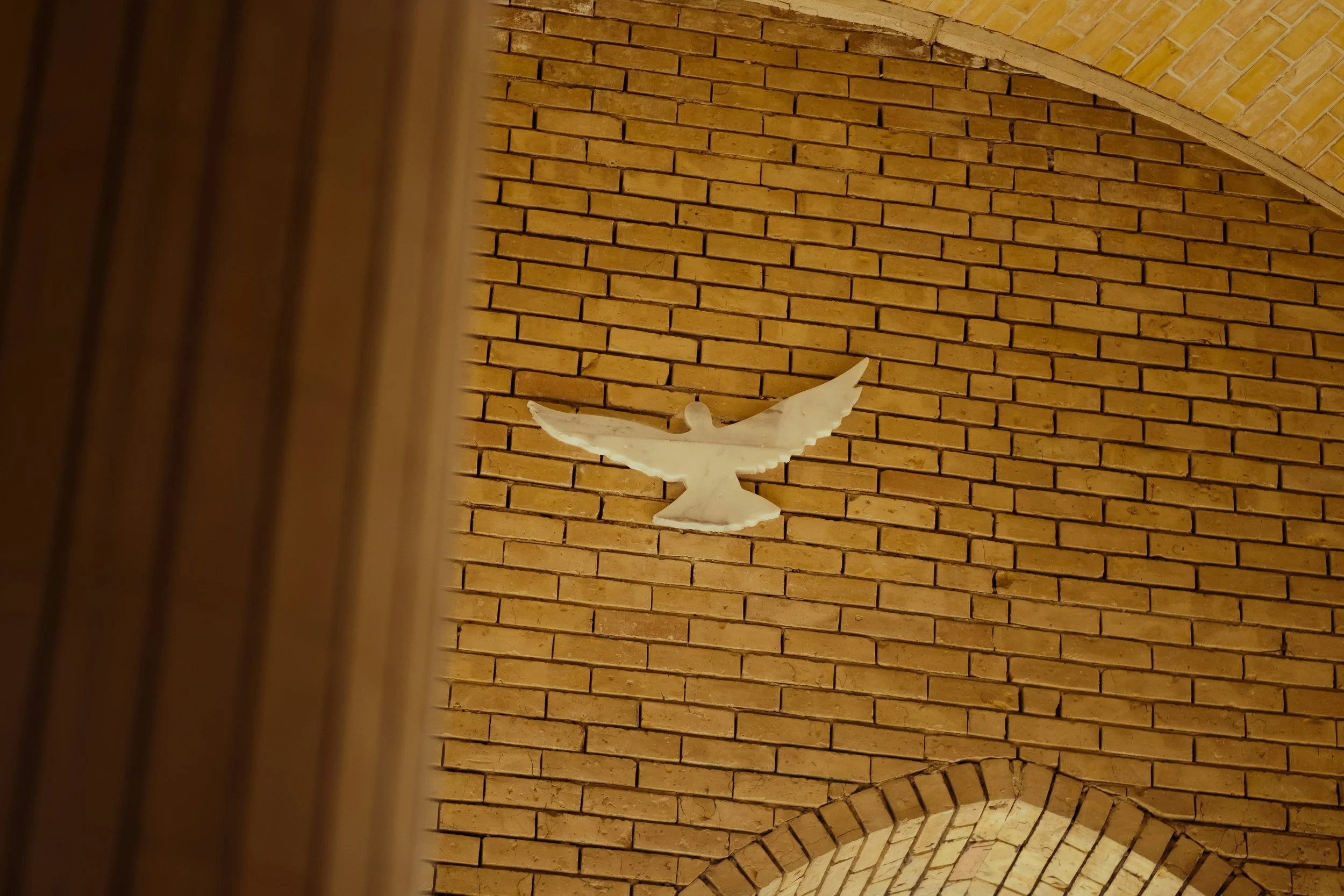 A white dove sculpture mounted on a brick wall in a space with an arched brick ceiling.