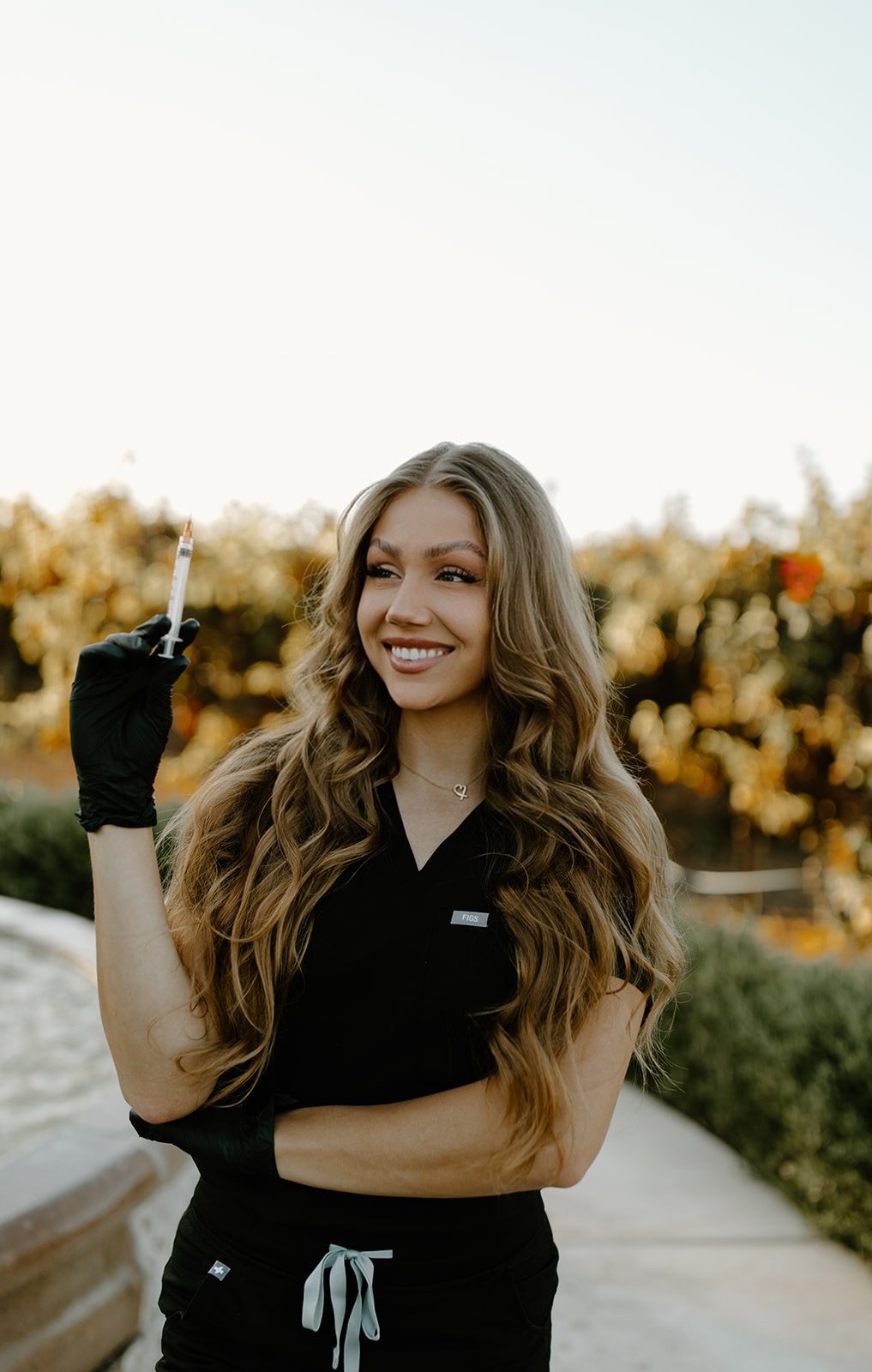 A young woman smiling outdoors holding a syringe in her gloved hand, wearing a black uniform with a name tag and a necklace, with trees in the background during sunset.