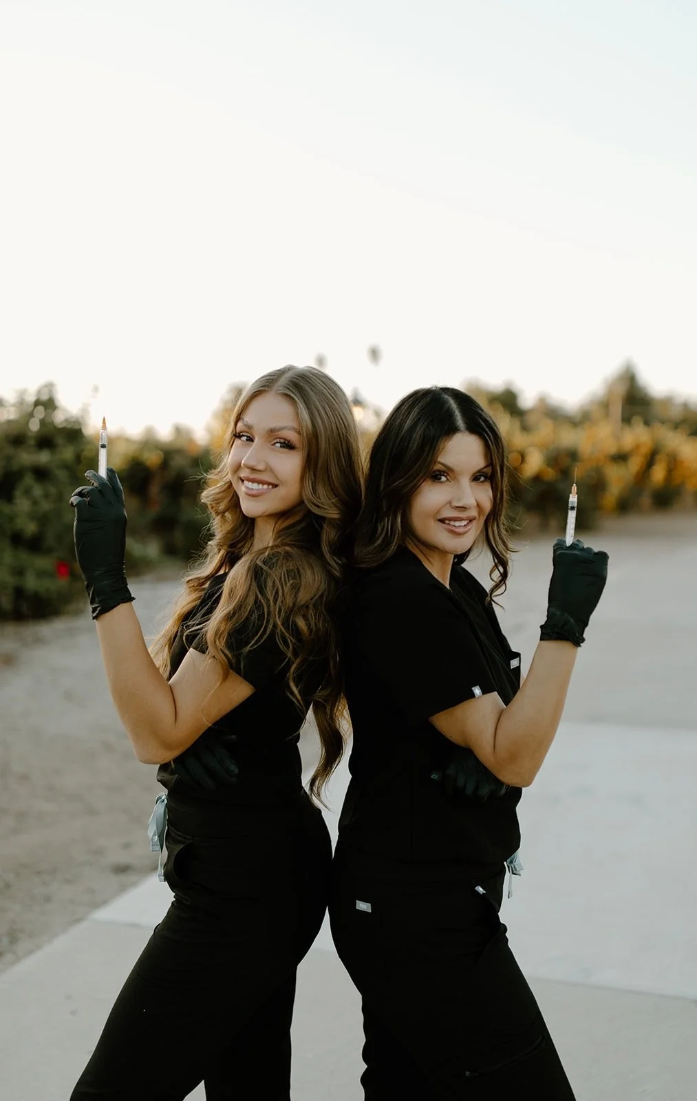 Two women in black scrubs and black gloves holding syringes, standing back-to-back outdoors during sunset, smiling and looking at the camera.
