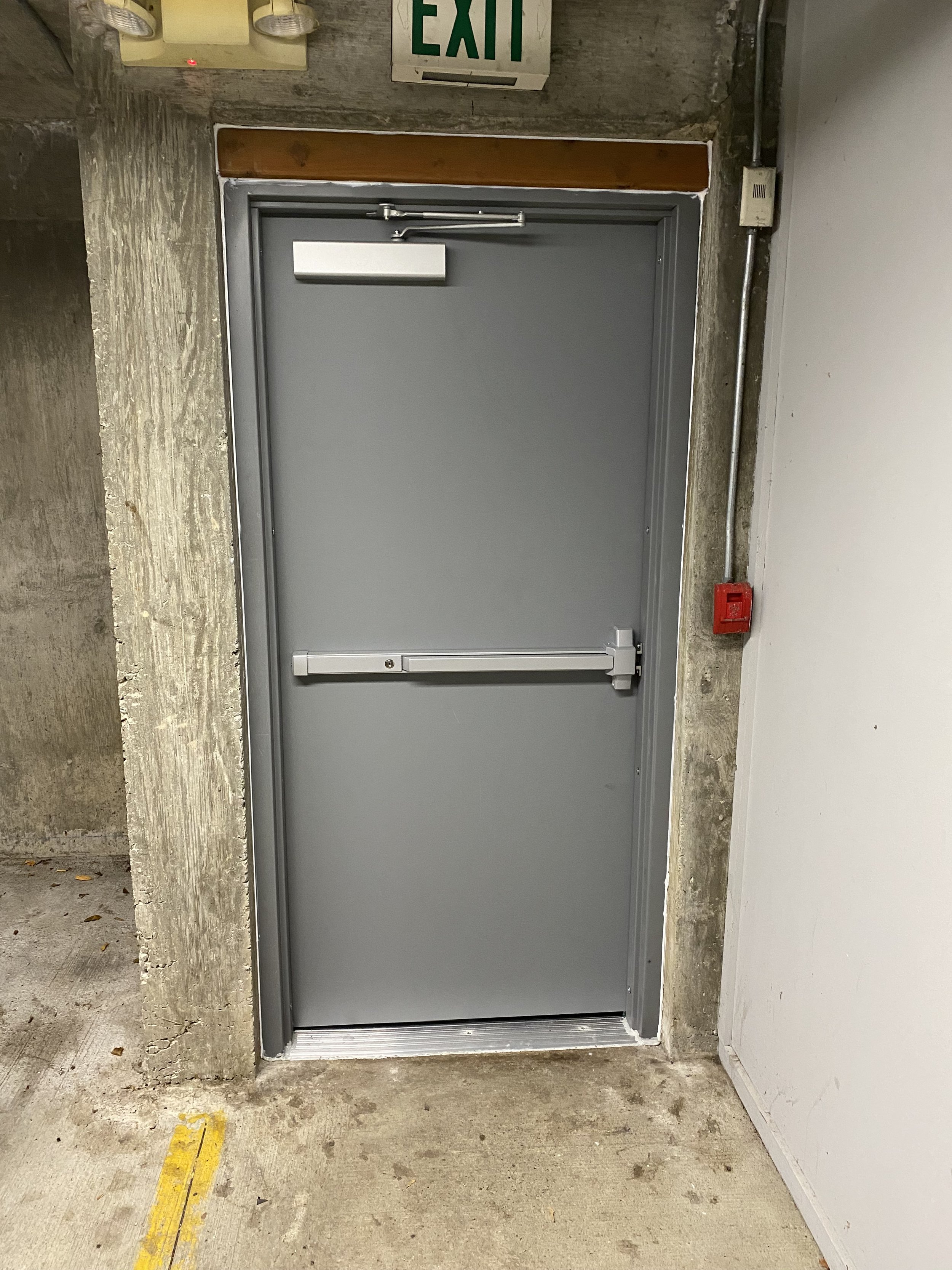 A gray metal door with a push bar, in a concrete wall with an exit sign above. There is a red fire alarm pull station on the right side of the door, and a small white box or sensor on the left side of the door. The floor is concrete with some dirt and debris.