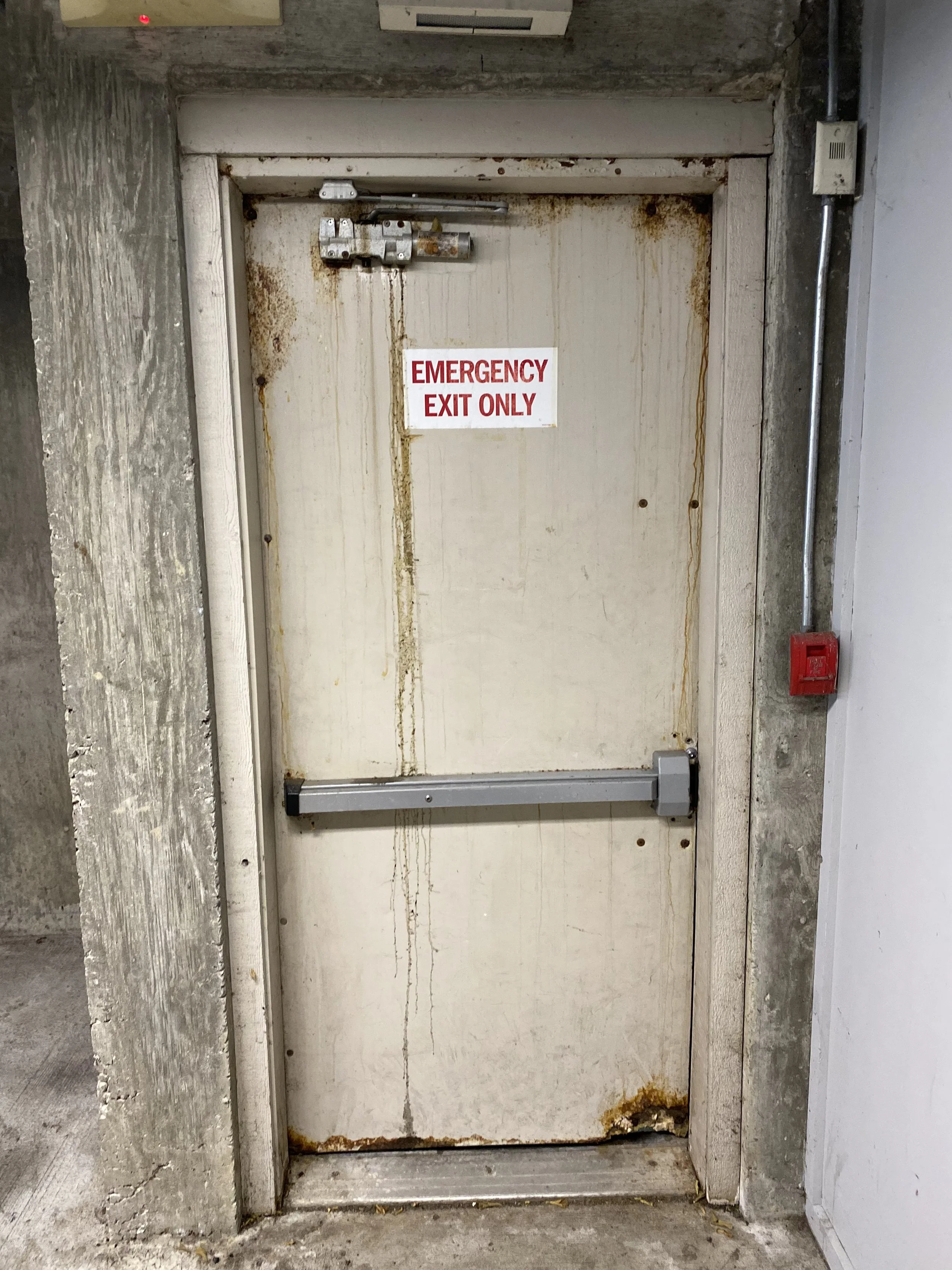 A rusted and worn metal emergency exit door with a sign that says 'EMERGENCY EXIT ONLY' in red letters. The door has a hydraulic door closer at the top, rust stains running down, and is set in a concrete wall with visible pipes and wiring to the right.