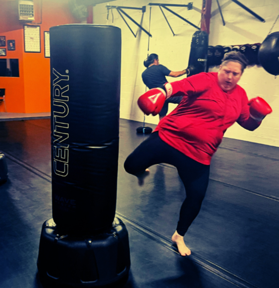Woman practicing kickboxing with red gloves and red shirt, kicking a punching bag at a gym.