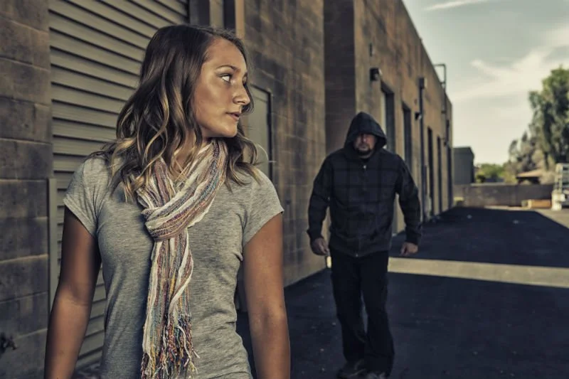 A woman with wavy hair and a scarf walking past a man in a hooded jacket on a city sidewalk.