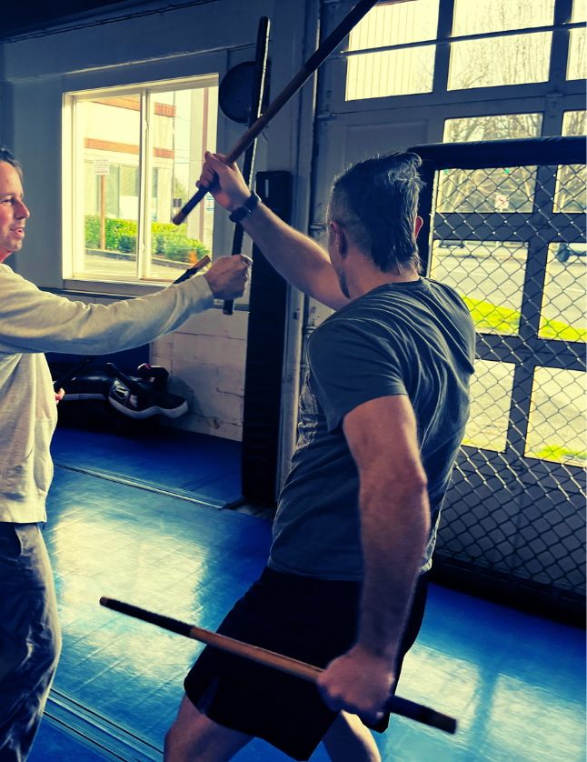 Two men practicing martial arts with staffs inside a gym, one is blocking the other's strike, with a chain-link door and windows showing trees outside.