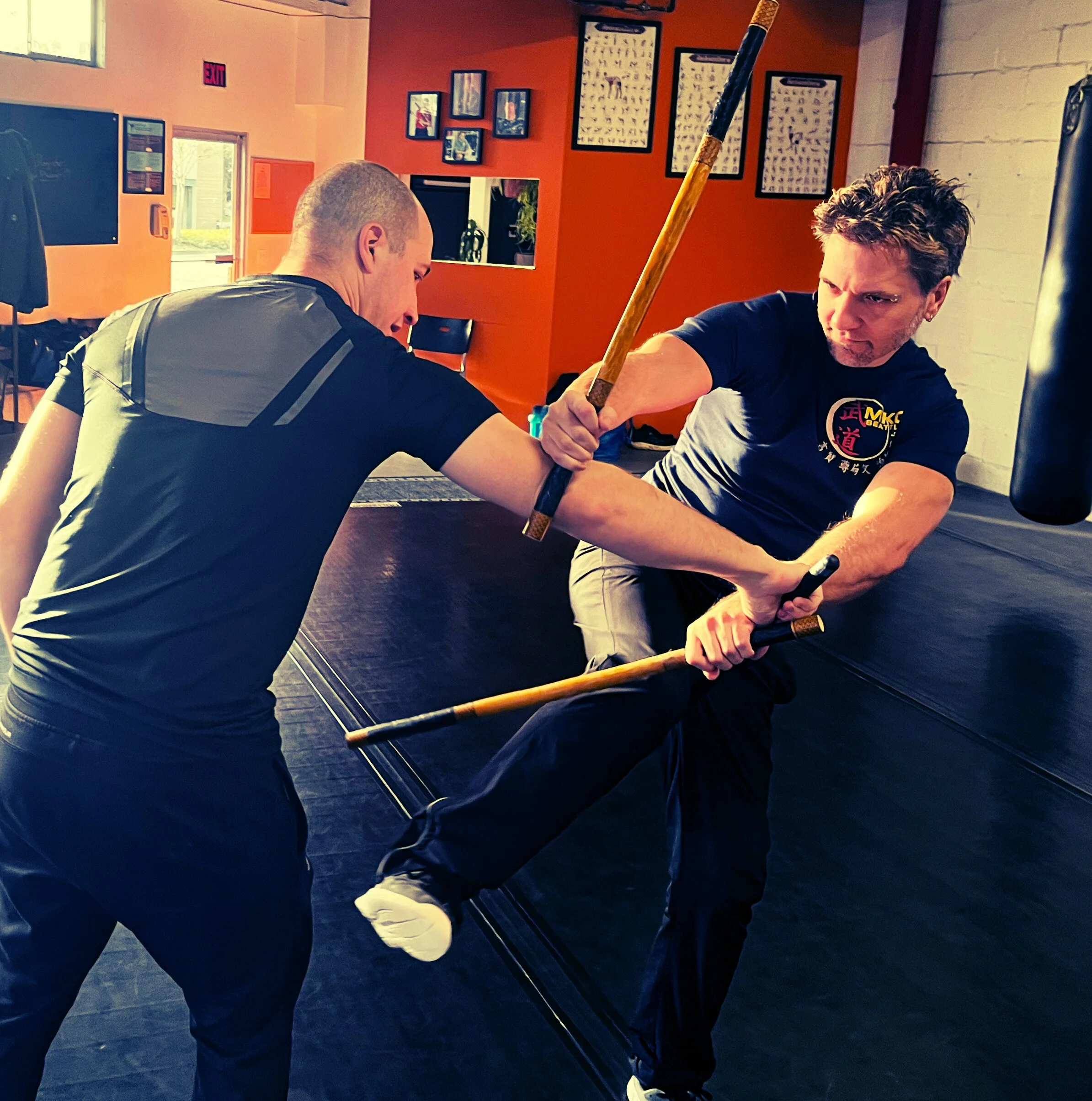 Two men engaging in a martial arts training exercise involving an arm lock with sticks in a gym setting, with orange and white walls decorated with framed pictures.