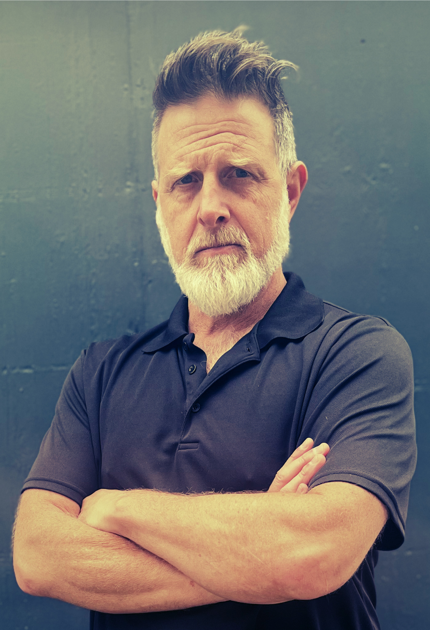 A middle-aged man with gray hair and beard, wearing a black polo shirt, standing with arms crossed in front of a dark gray wall.