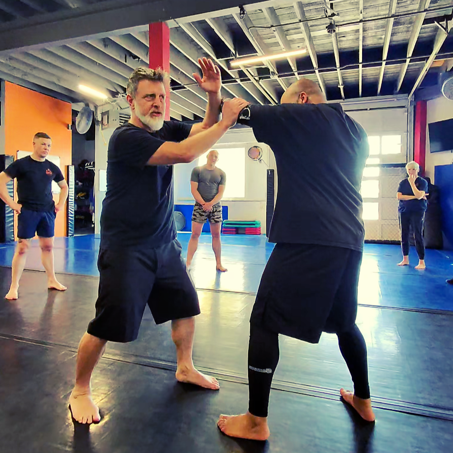 Two men practicing martial arts or self-defense techniques in a gym with a blue mat, while three women observe in the background.