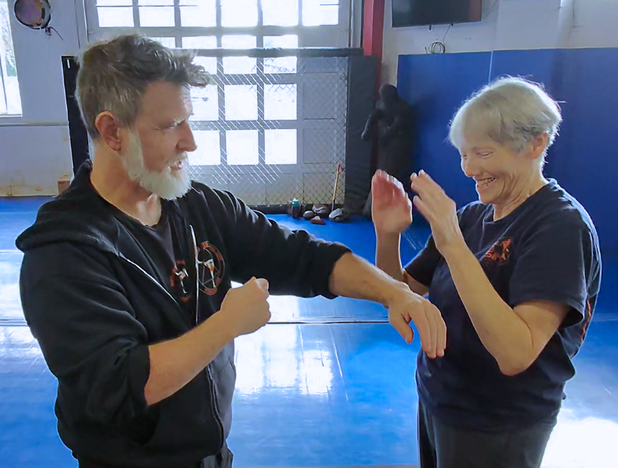Two people practicing martial arts in a gym, one with a gray beard and the other with short blonde hair. They are smiling, and the person with a gray beard is demonstrating a technique on the other.