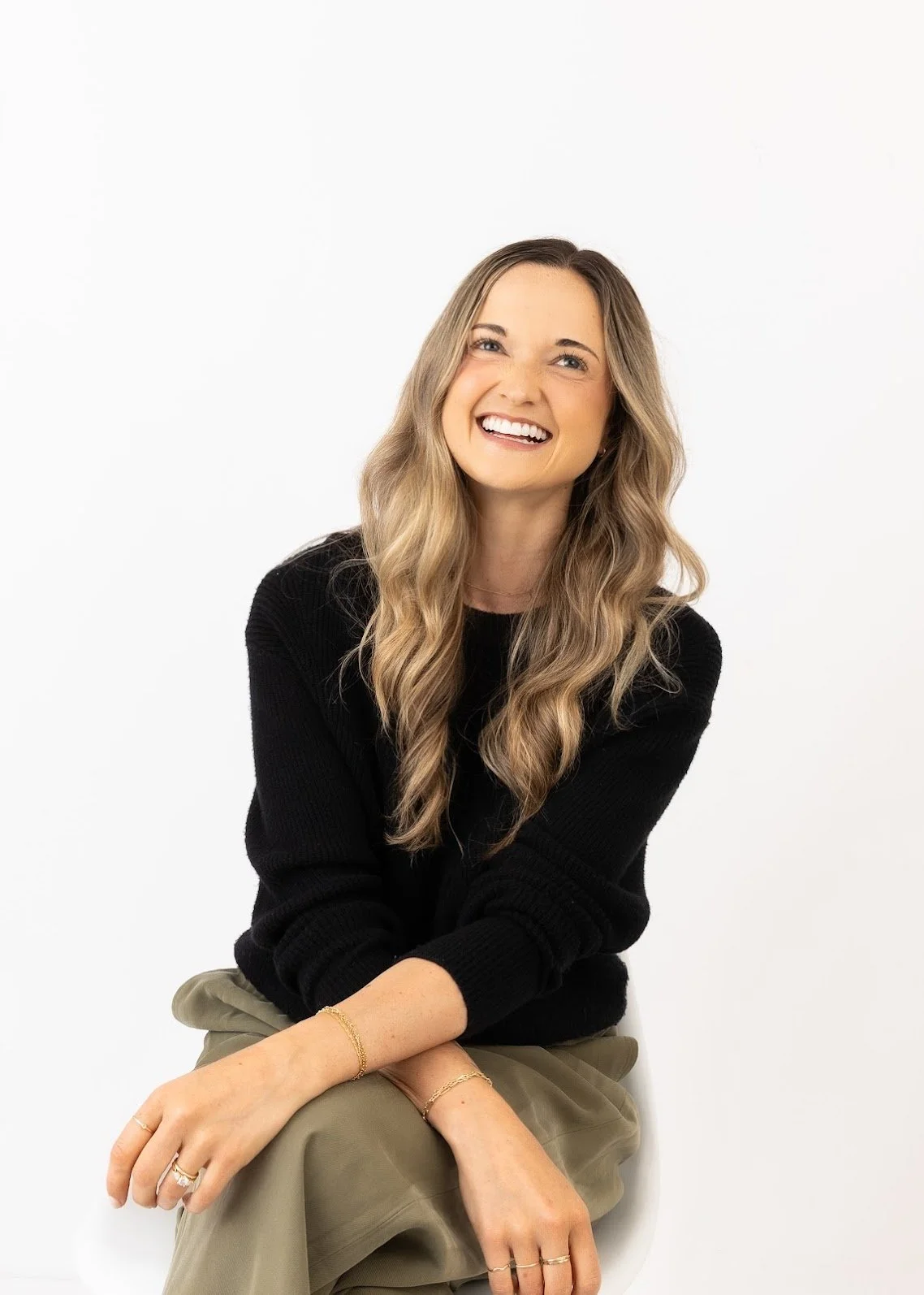 A woman with long wavy blonde hair, wearing a black sweater and beige pants, sitting against a white background, smiling and looking slightly to the side.