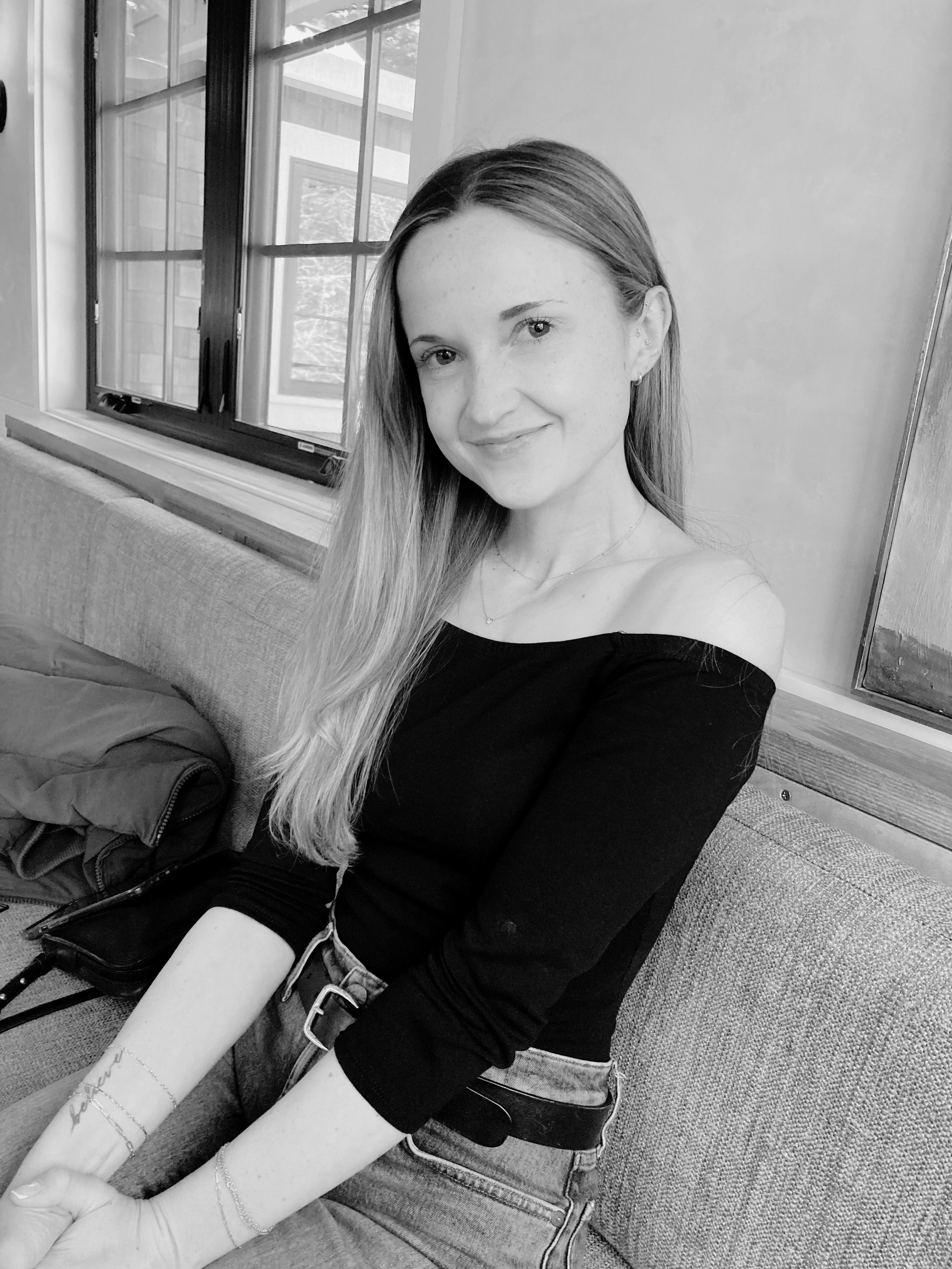 A woman with long hair and earrings sitting on a couch near a window, smiling at the camera, in black and white.