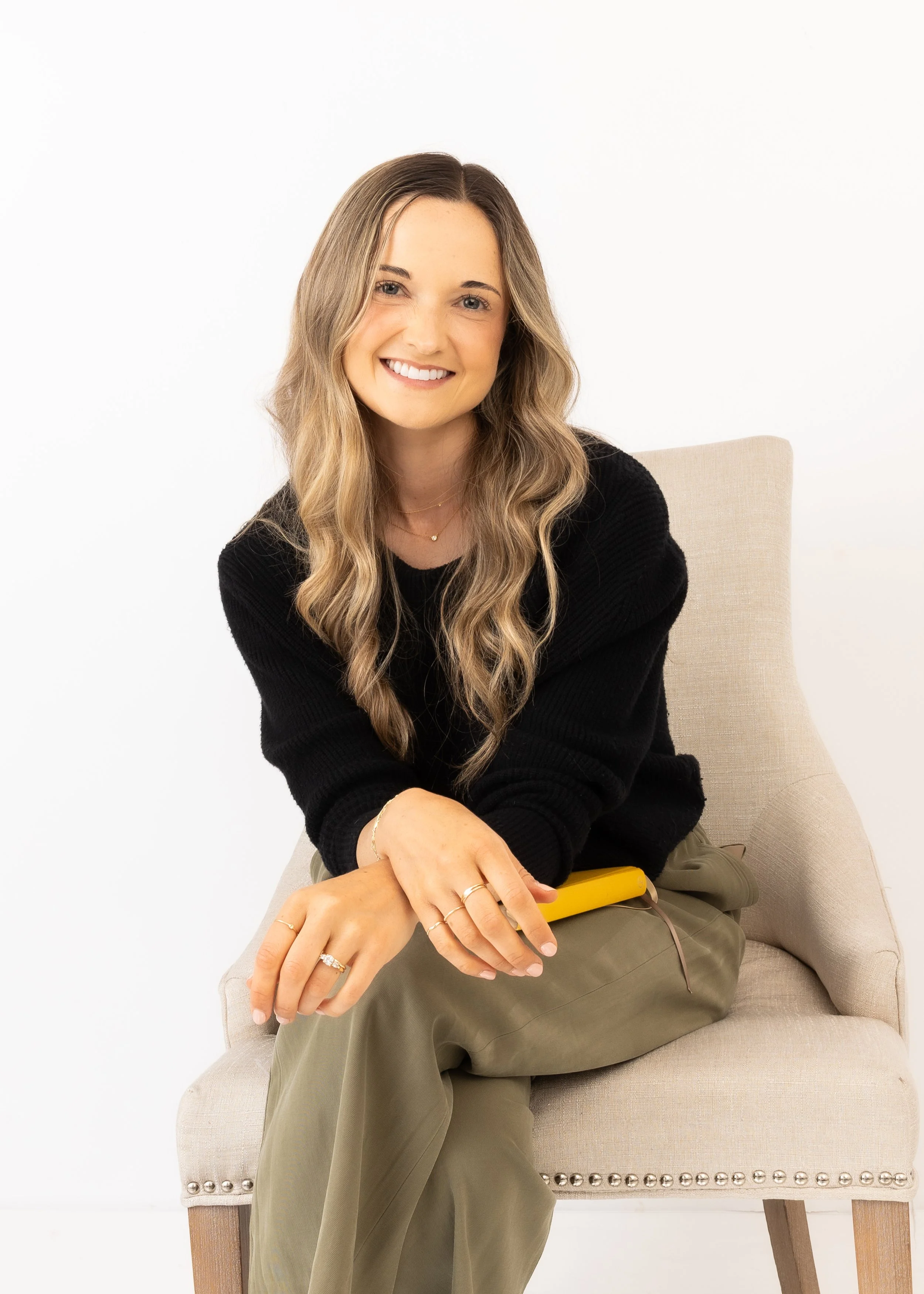A woman with wavy blonde hair and a black sweater sitting on a beige chair with nailhead trim, smiling at the camera, holding a yellow device, dressed in khaki pants and wearing jewelry against a plain white background.