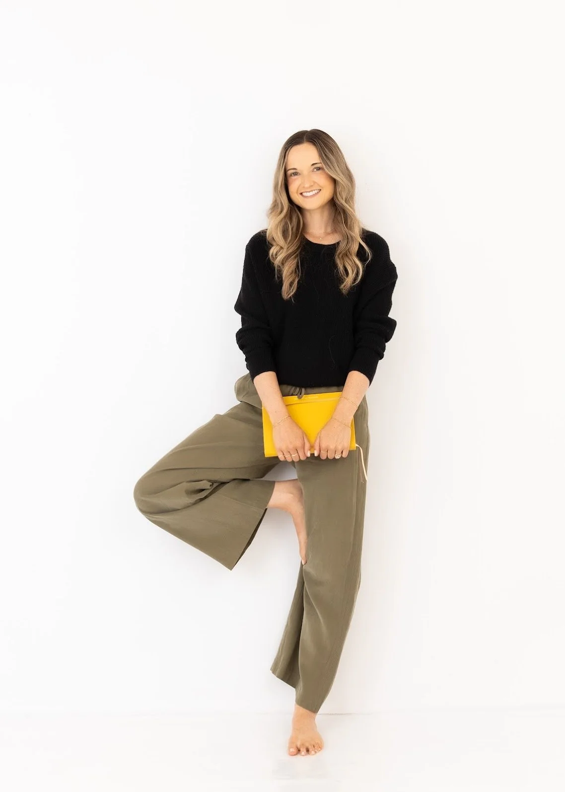 A woman with long, wavy hair, smiling, holding a yellow notebook, standing against a white wall, with one foot on the ground and the other foot resting on her knee.