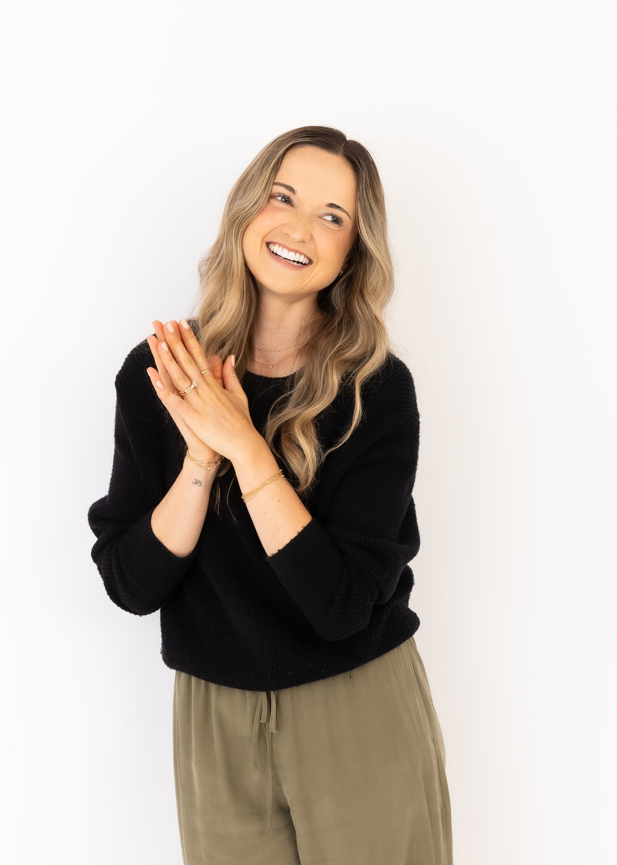 A woman with long wavy blonde hair, smiling and looking to her left, wearing a black sweater and beige pants, standing against a plain white background.