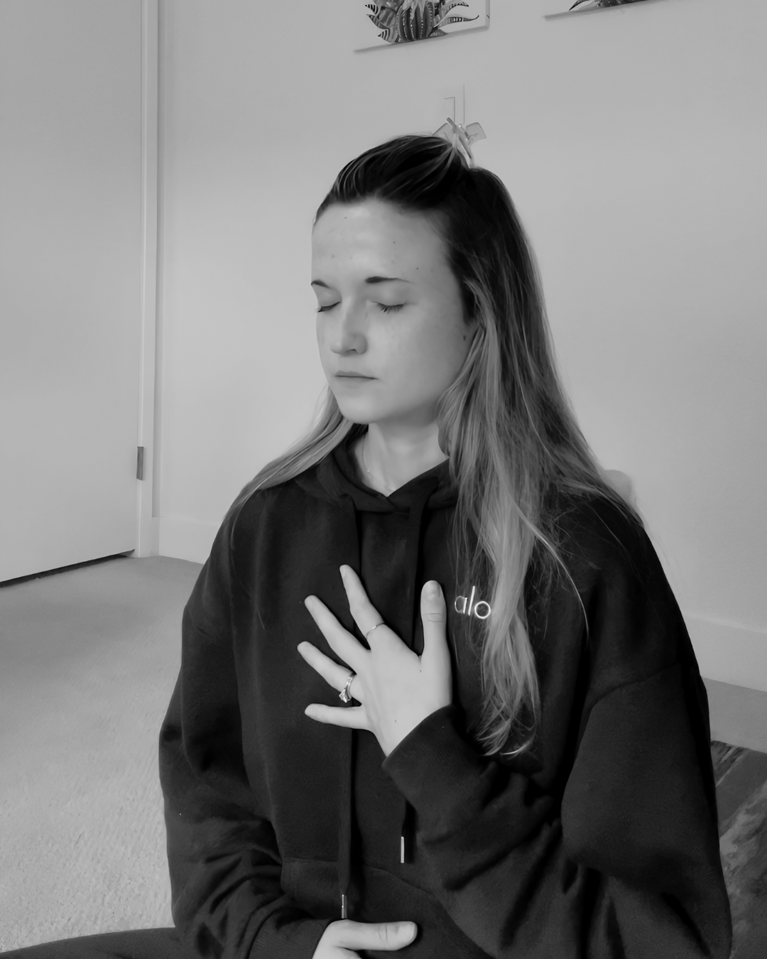 Black and white photo of a woman with closed eyes, wearing a dark hoodie with 'alo' written on it, holding her hand on her chest in a calm pose.