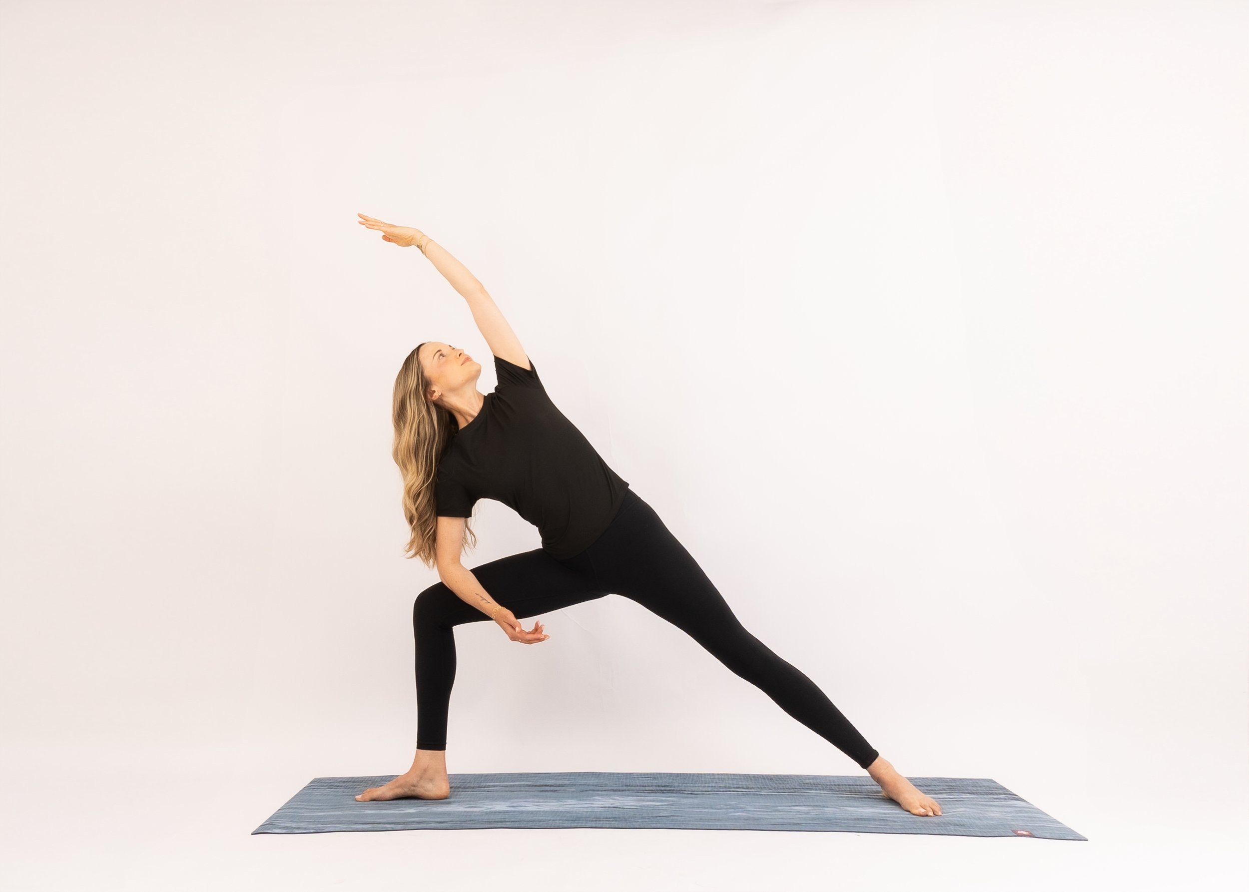 A woman practicing yoga on a mat, in a side lunge pose with one hand reaching toward the ceiling and the other hand resting on her side, against a plain white background.