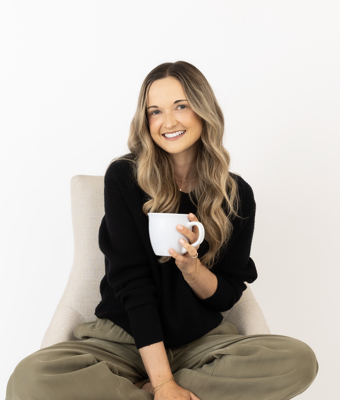 A woman with long, wavy blonde hair smiling and holding a white mug, sitting cross-legged in a beige chair against a white background.