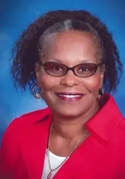 A woman with glasses, curly gray and black hair, smiling, wearing a red blazer and a white top, against a blue background.