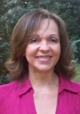 A woman with shoulder-length brown hair, smiling, wearing a pink collared shirt, standing outdoors with trees in the background.