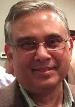 A man with glasses and short gray hair smiling, wearing a collared shirt and blazer, indoors.