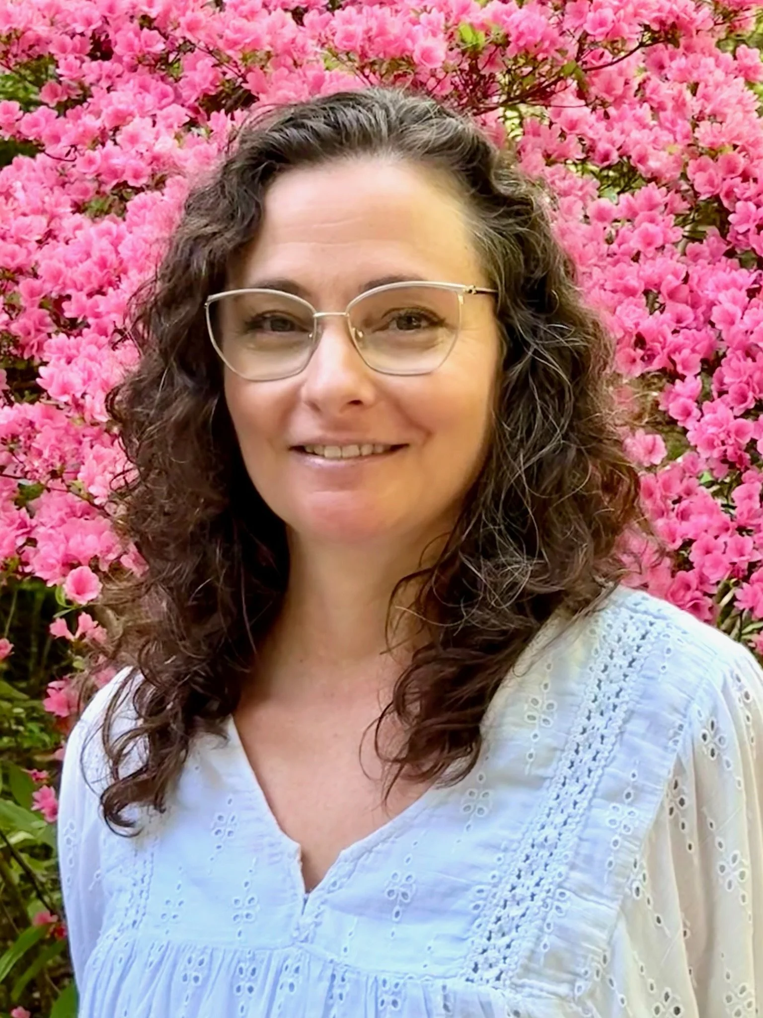A woman with curly brown hair and glasses smiling, standing in front of a pink flowering bush, wearing a white eyelet blouse.