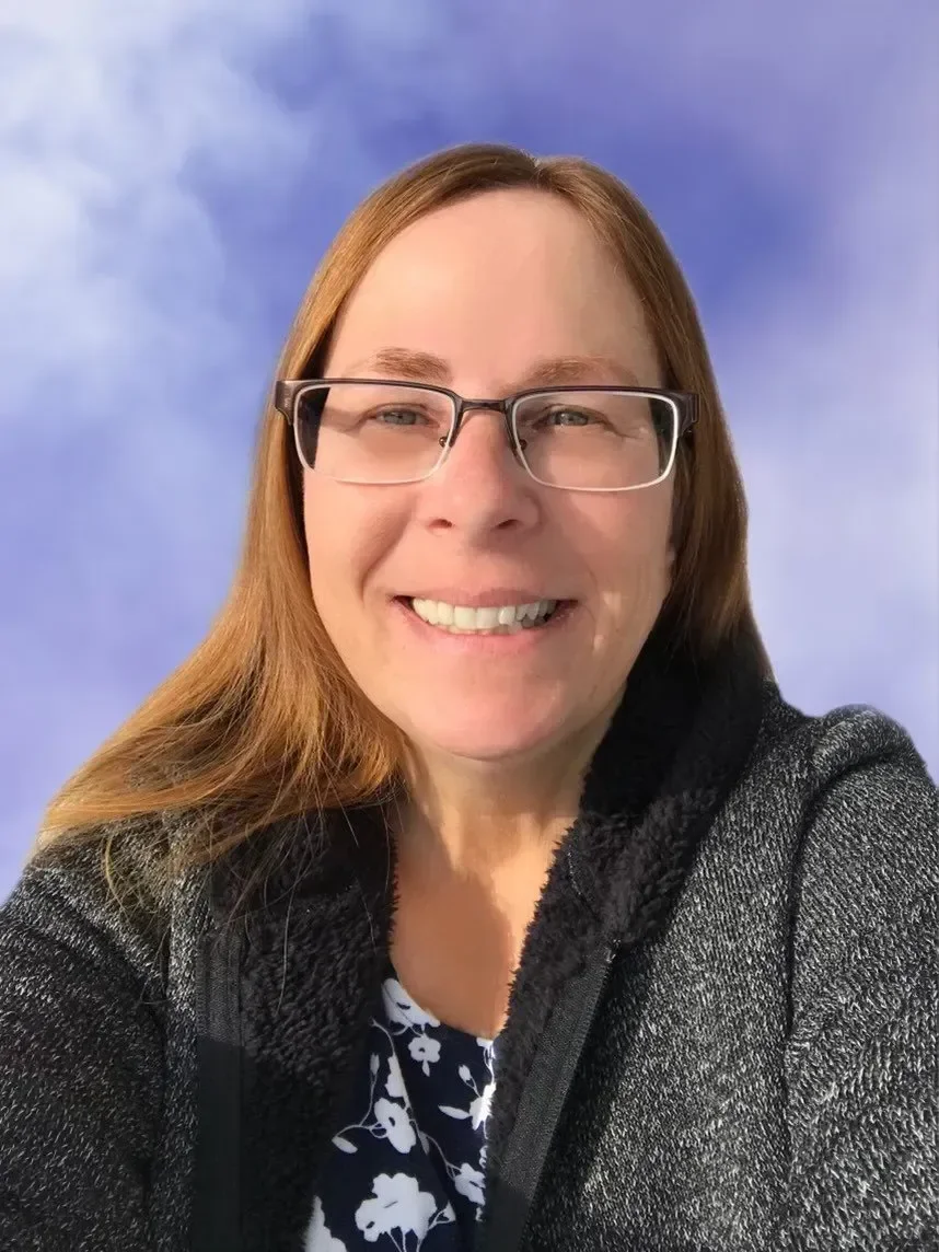 A woman with long reddish-brown hair and glasses smiling in front of a blue sky background.