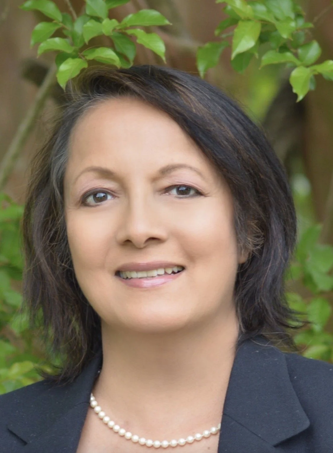 A woman with short dark hair, light makeup, wearing a pearl necklace and a black blazer, smiling outdoors with green leaves in the background.