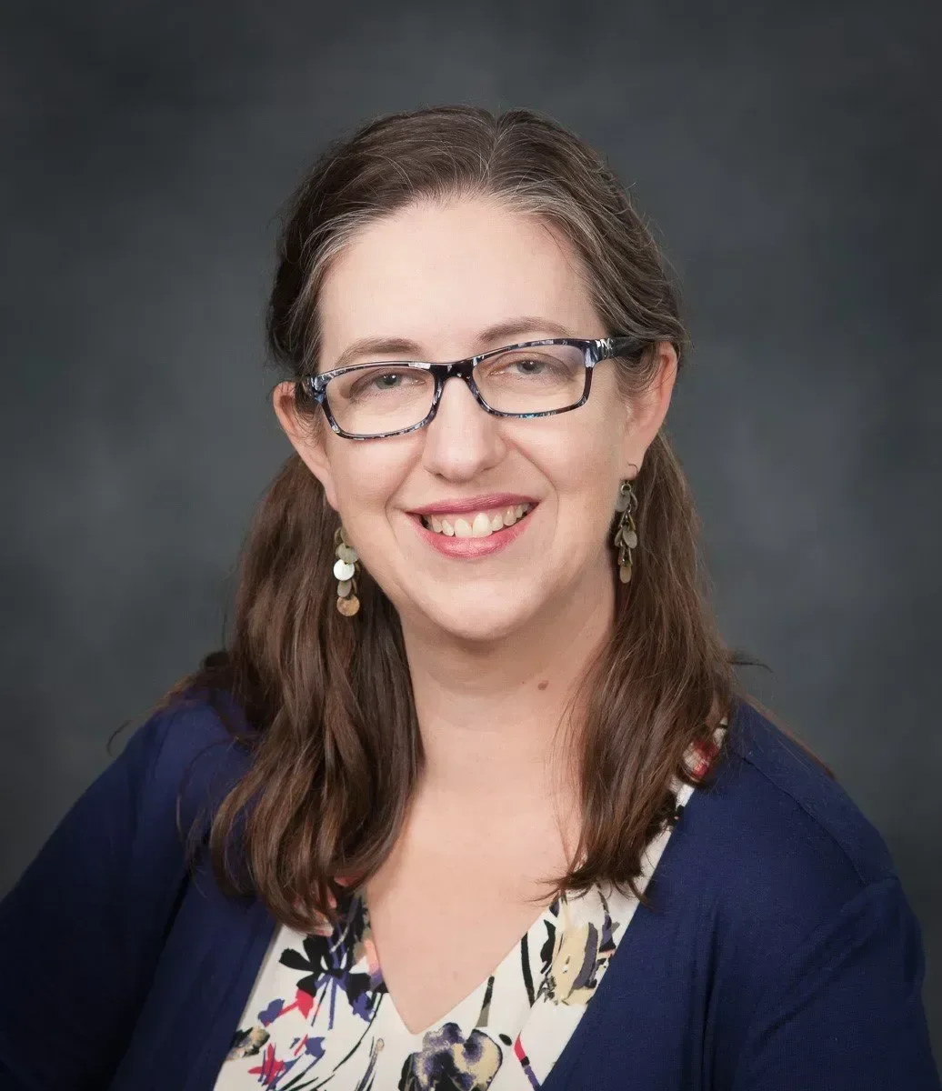 A woman with brown hair, glasses, and earrings smiling in front of a dark gray background.