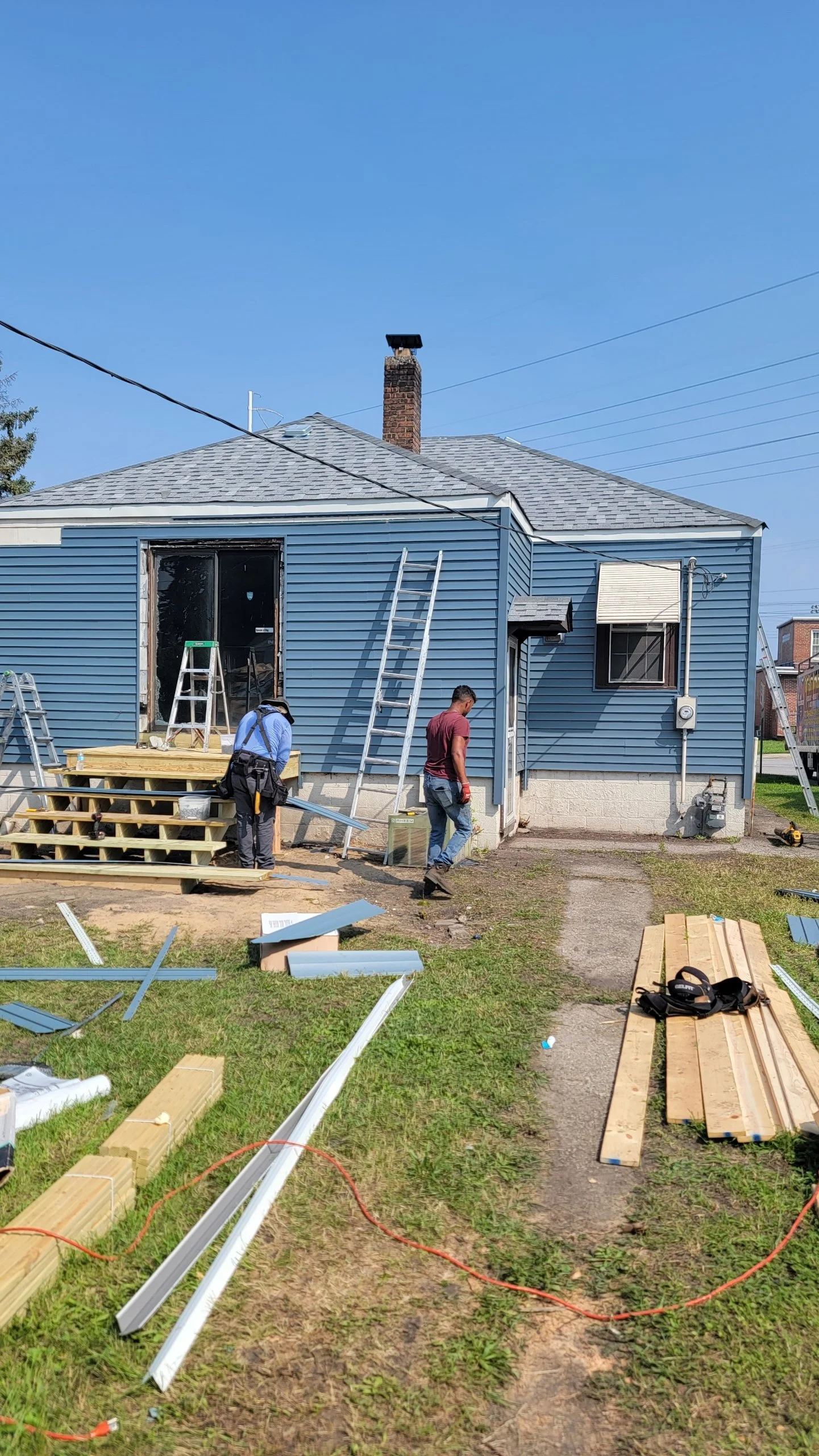 House renovation with workers installing siding. Construction tools and materials scattered on the yard, including wooden planks, metal strips, and ladders.