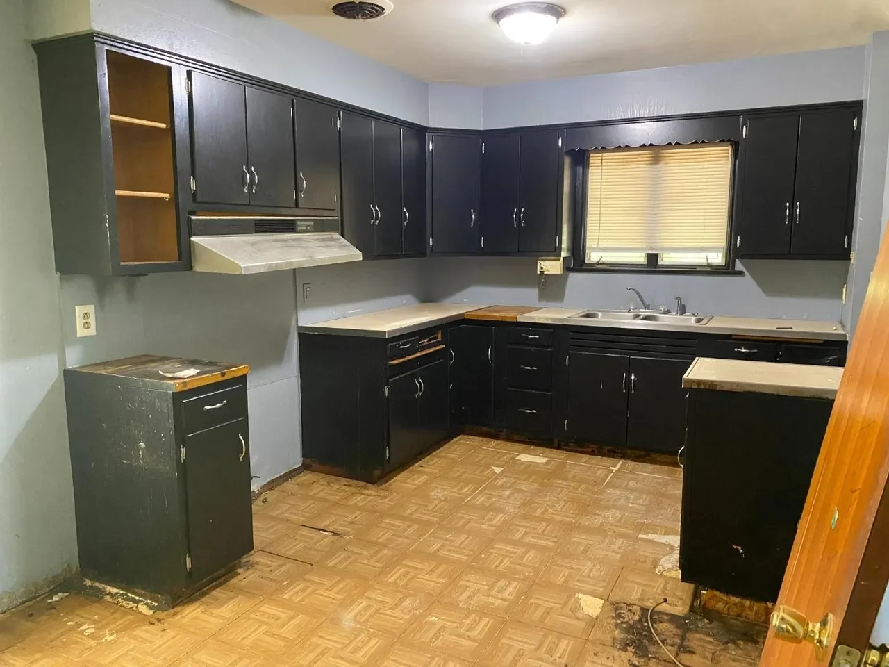 Kitchen with black cabinets, a window with blinds, and a worn wooden floor. Some cabinets and counter space appear damaged or in disrepair.